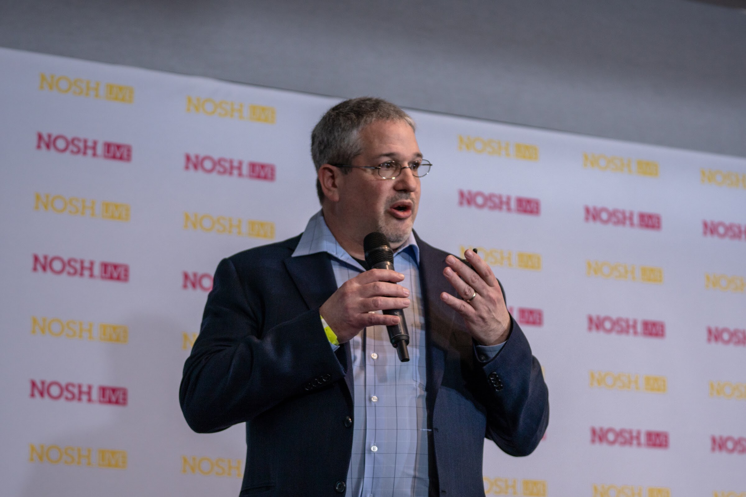Man with glasses and grey hair in a navy blazer and light blue shirt speaking into a microphone in front of a repeating pink and yellow Nosh Live backdrop.