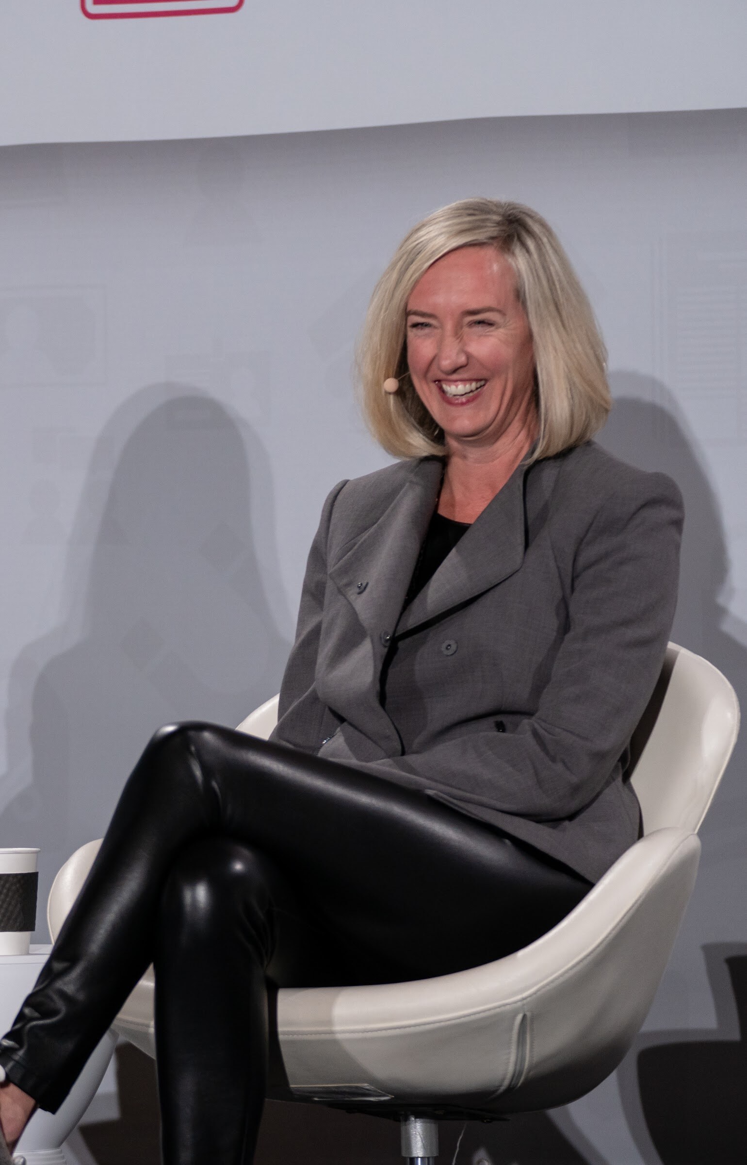 Woman with shoulder-length blonde hair in a grey blazer and black leather pants laughing in a white chair on stage.