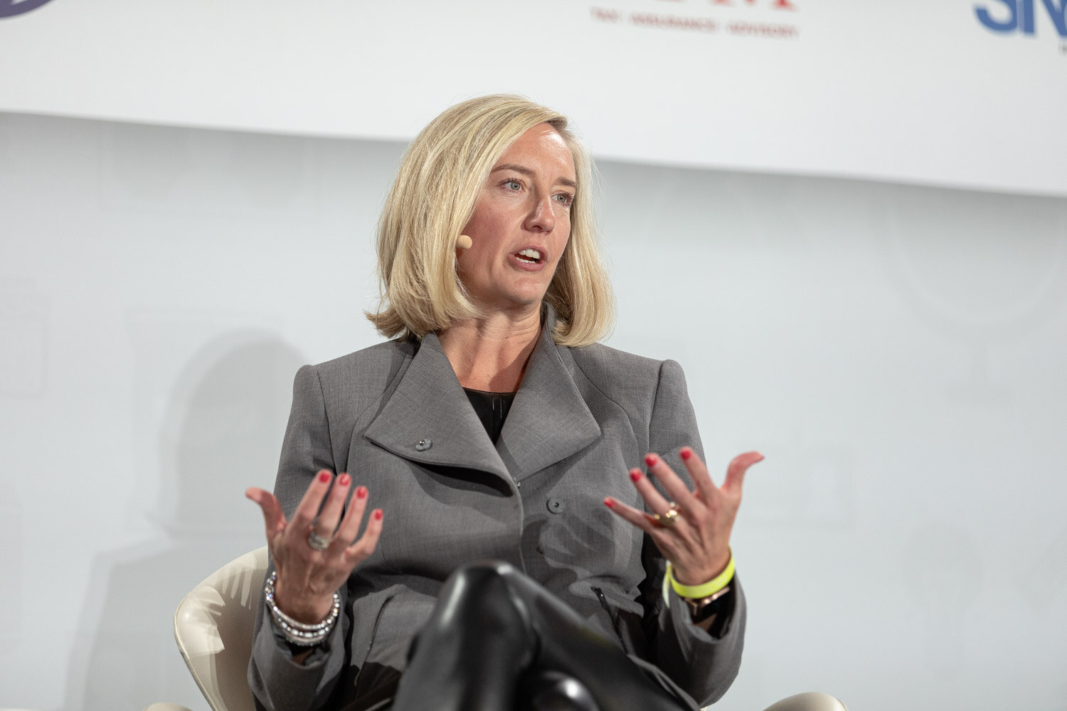 Woman with blonde bob in a grey blazer and black leather pants, with a yellow wristband, gesturing with both hands while seated on stage.