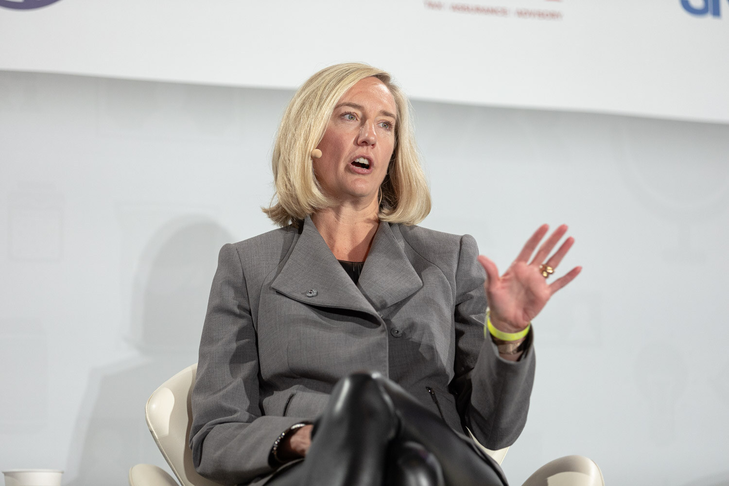 Woman with blonde bob in a grey blazer and a headset microphone with one hand raised, seated in a white chair in front of a white backdrop.