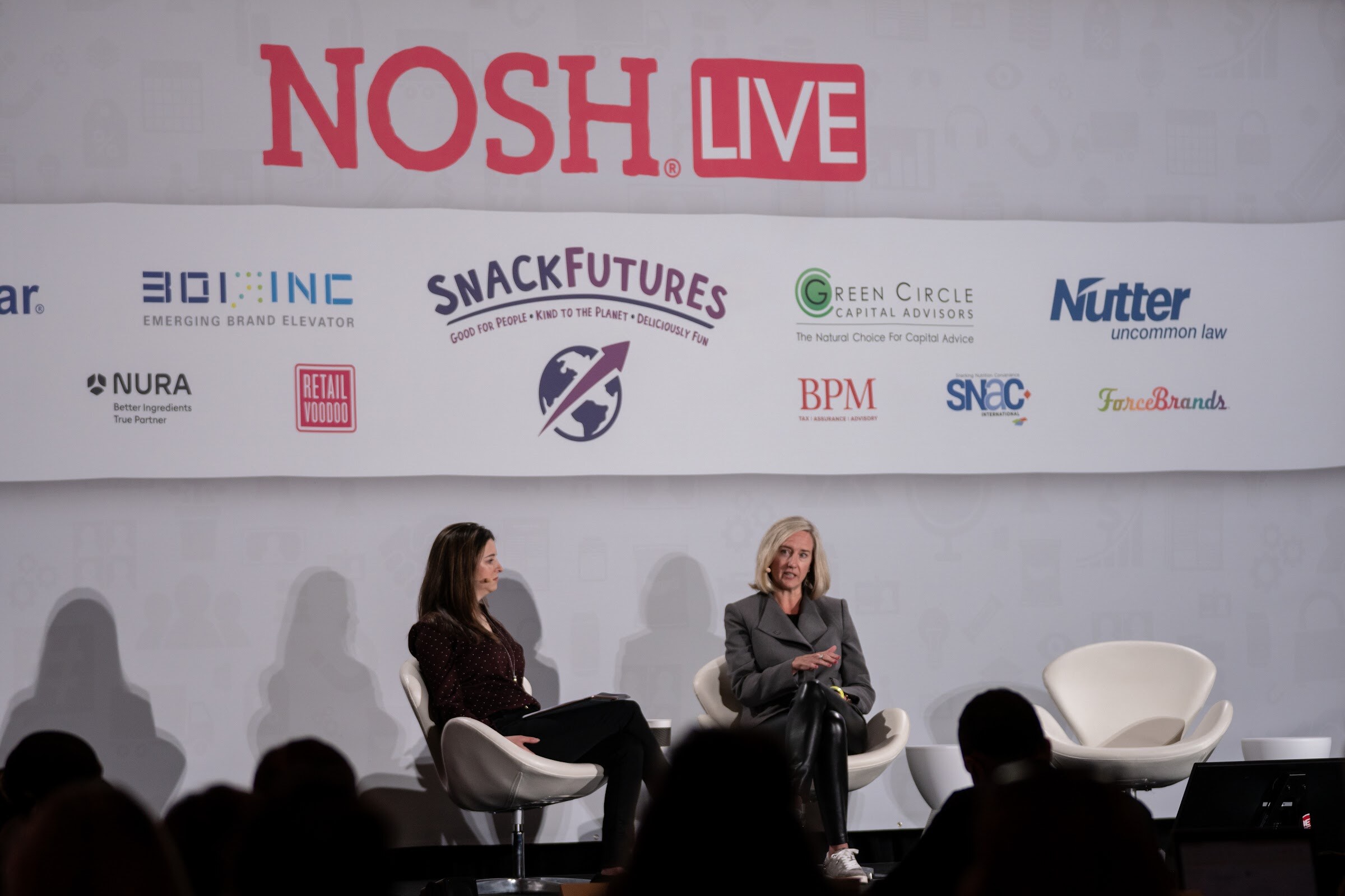 Wide view of two women seated in white chairs on stage in front of a Nosh Live banner with sponsor logos including 301 Inc, SnackFutures, Green Circle Capital Advisors and Nutter, with the audience in silhouette.
