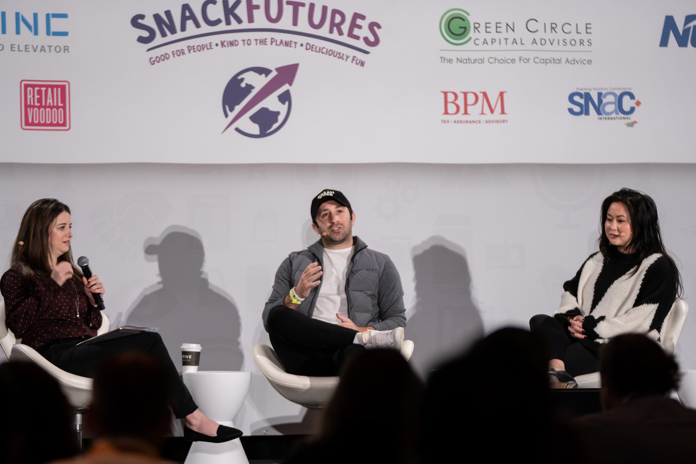 Woman in a burgundy polka-dot top holding a microphone, a man in a black cap and grey puffer vest and a woman in a black and white knit sweater seated on stage in front of SnackFutures and Green Circle Capital Advisors logos.