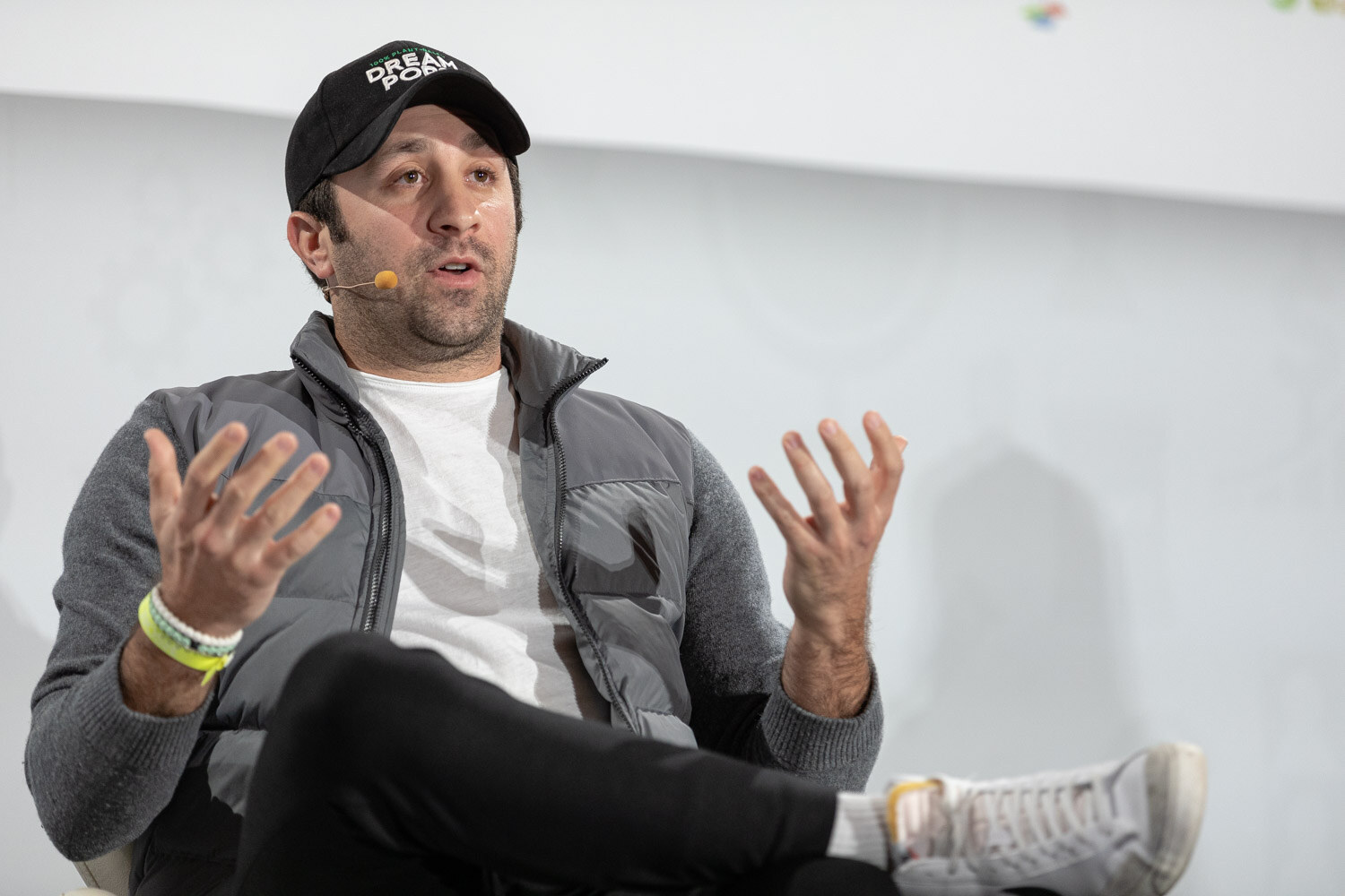 Man in a black Dream Pops cap, grey puffer vest and white t-shirt with both hands raised, seated and talking, with yellow and white wristbands.