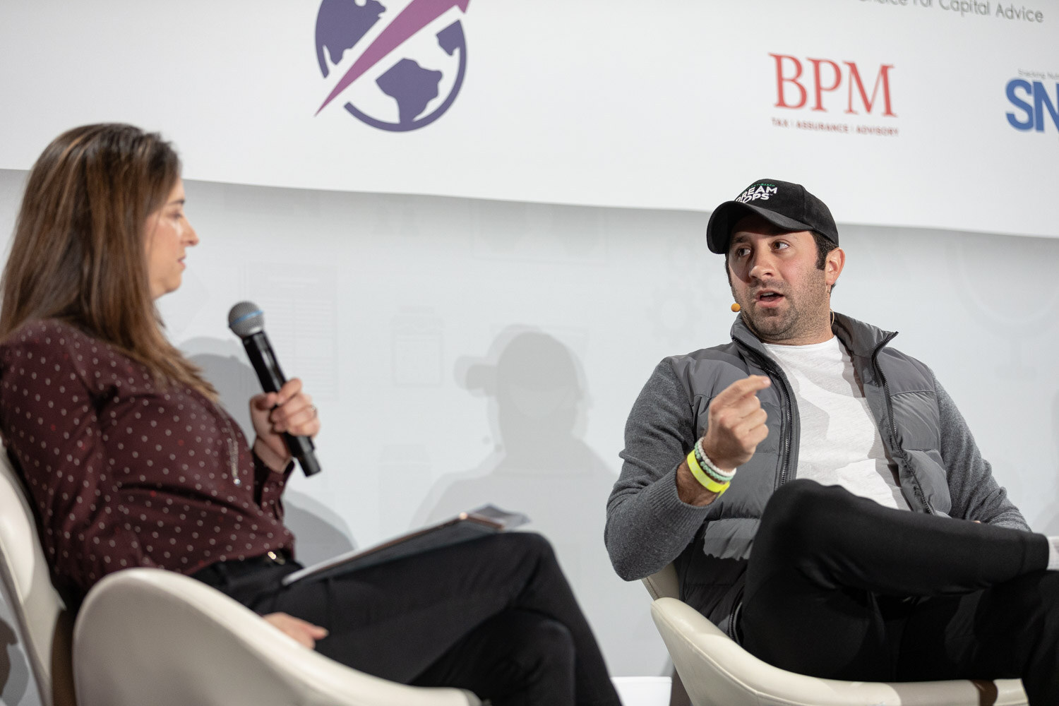 Woman in a burgundy polka-dot top holding a microphone at the left and a man in a black Dream Pops cap and grey vest gesturing at the right, in front of BPM and SNAC logos.