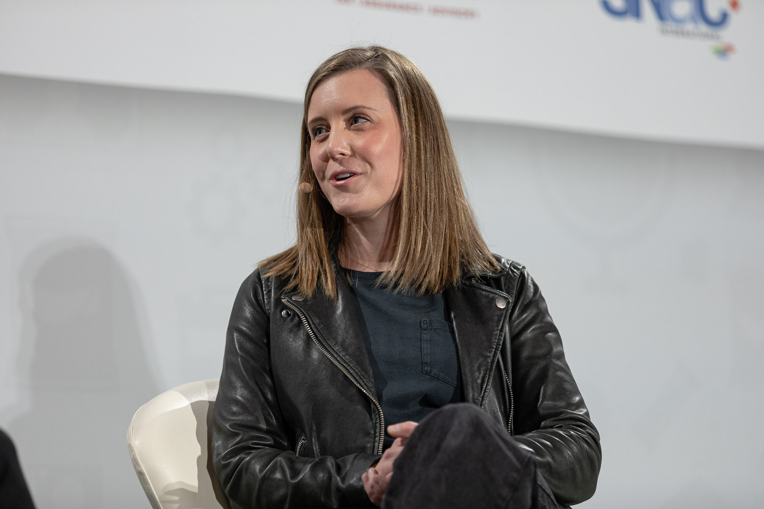 Woman with straight shoulder-length brown hair in a black leather jacket over a dark top seated in a white chair with her hands in her lap.