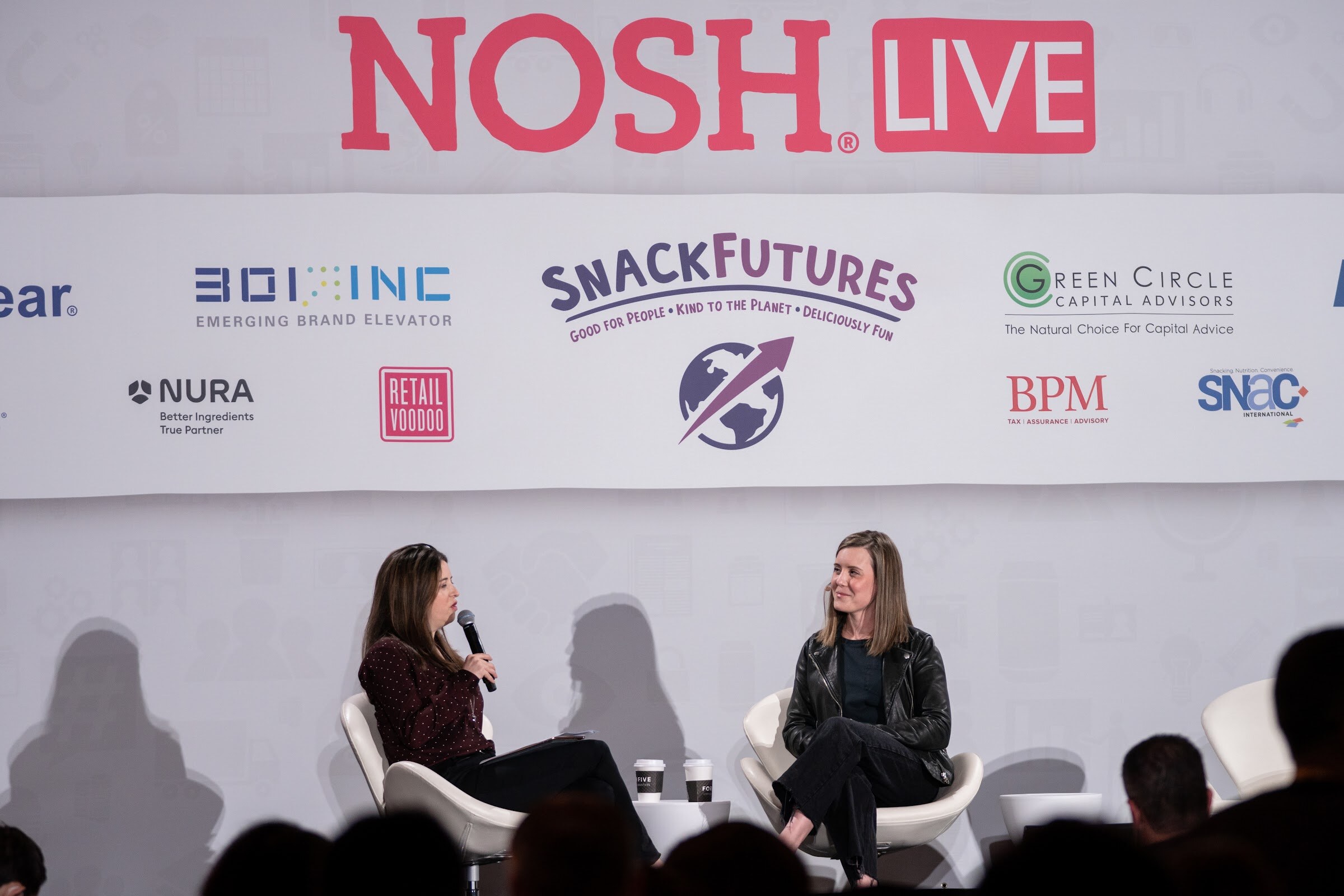Wide view of two women seated in white chairs on stage in front of a Nosh Live banner with sponsor logos including 301 Inc, SnackFutures and NURA, with the audience in silhouette.