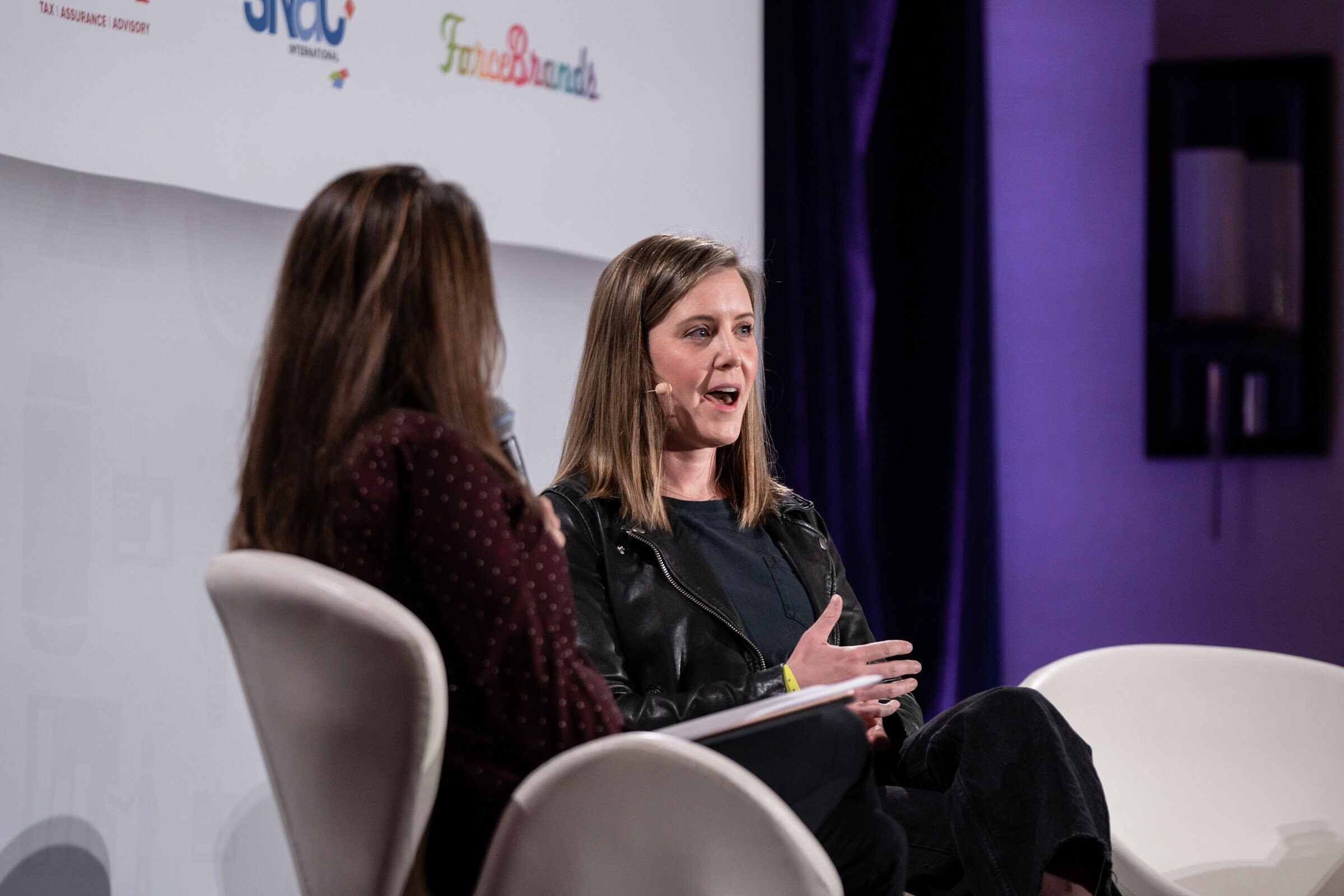Woman with shoulder-length brown hair in a black leather jacket gesturing while speaking, with the back of the moderator's head in the left foreground and a purple curtain behind.