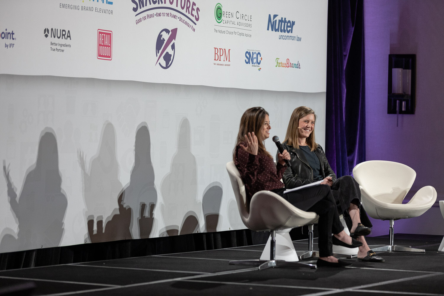 Two women seated side by side on white chairs on stage, one holding a microphone and gesturing, in front of a sponsor wall with NURA, Retail Voodoo, SnackFutures and Nutter logos.