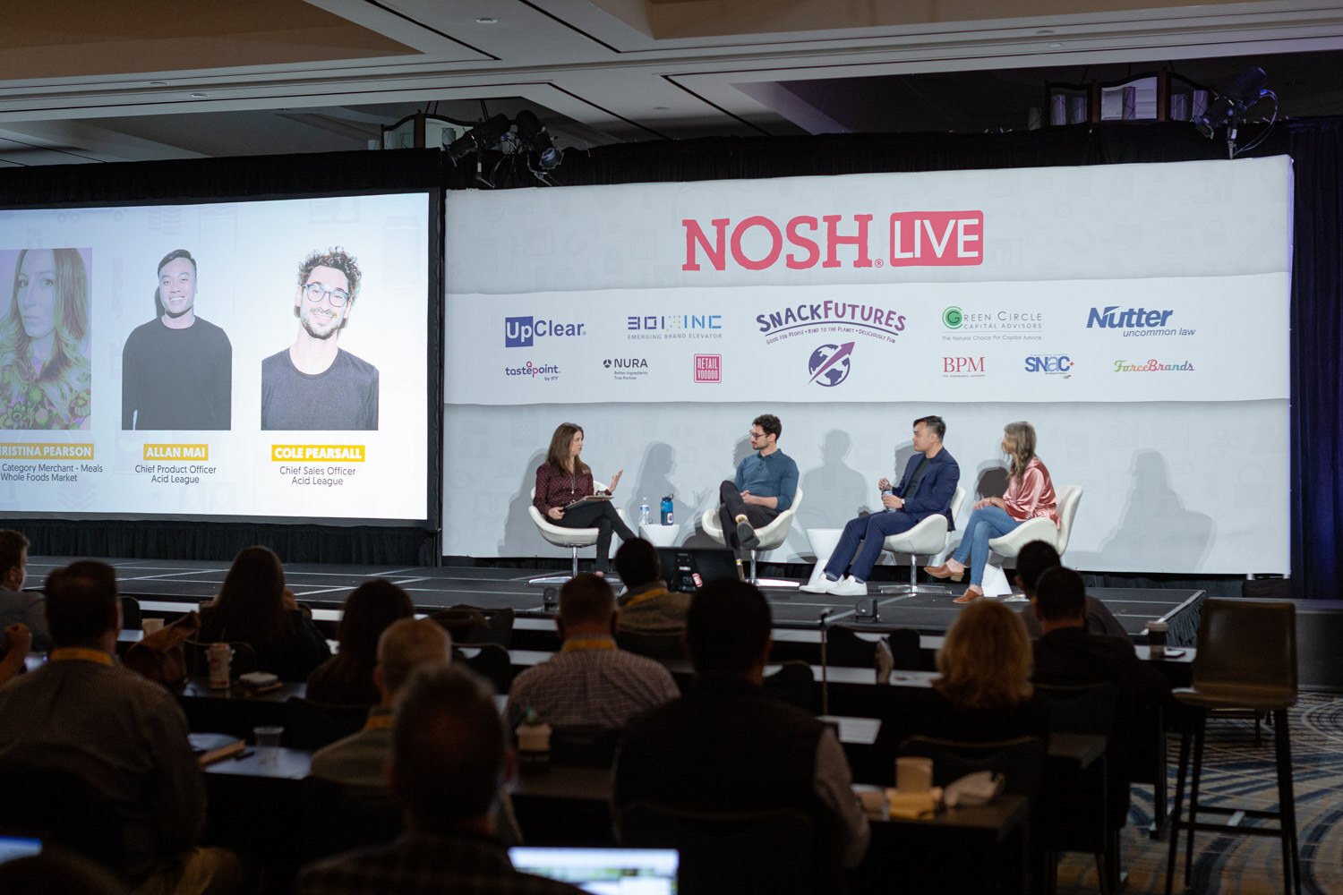 Wide view of a ballroom with four people seated on stage in front of a Nosh Live banner, a screen showing three speaker portraits with names and titles at the left and audience members at tables in the foreground.