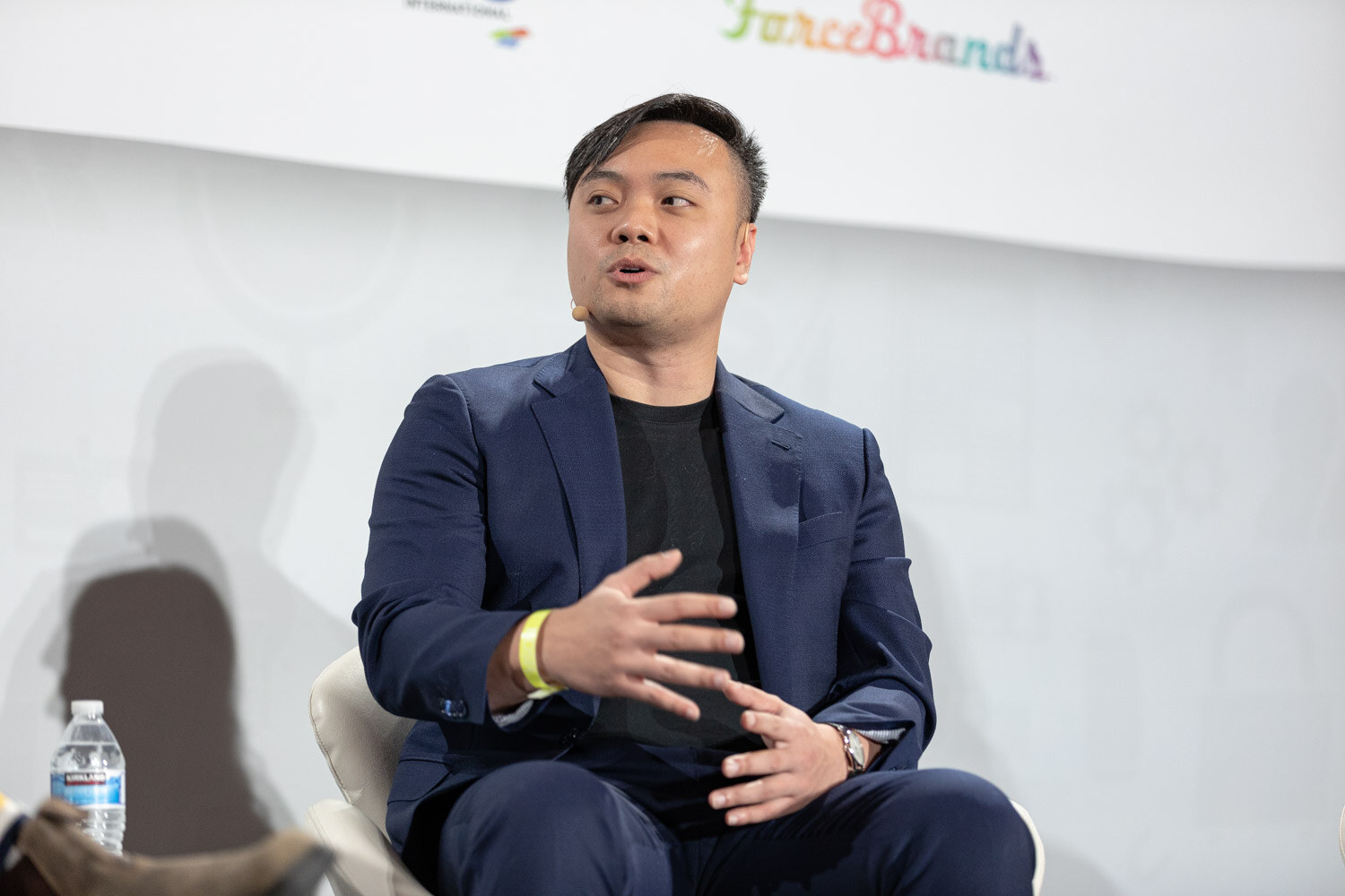 Man in a navy blazer over a black t-shirt gesturing with both hands, with a yellow wristband and a water bottle at his side, in front of a white backdrop.
