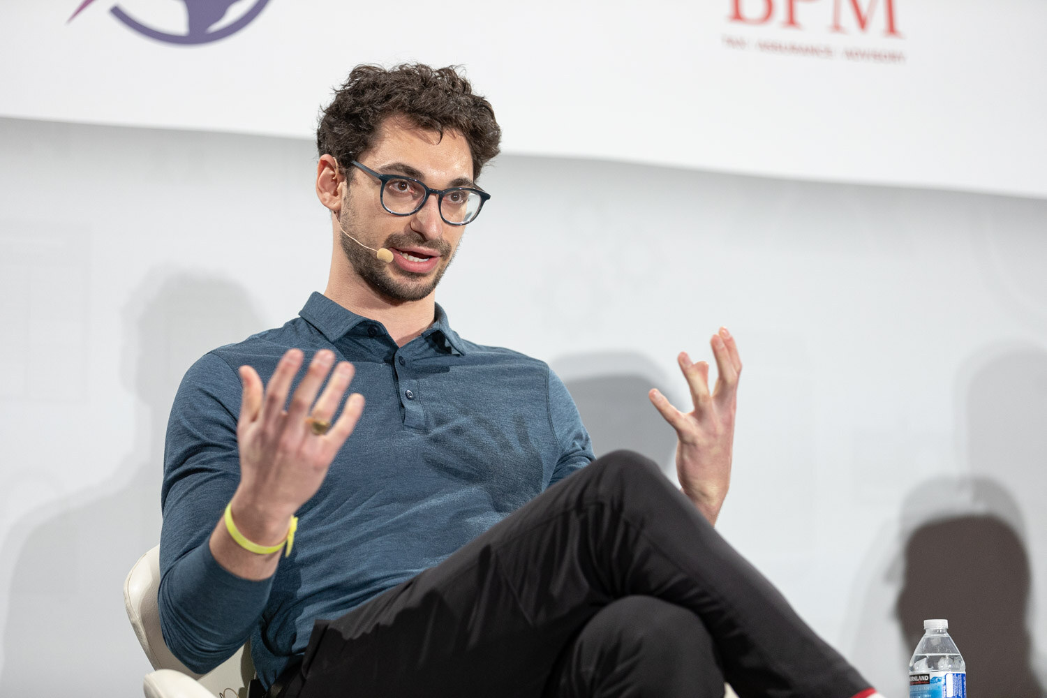 Man with dark curly hair, glasses and a beard in a blue polo shirt, with both hands raised and a yellow wristband, seated in front of a BPM logo.