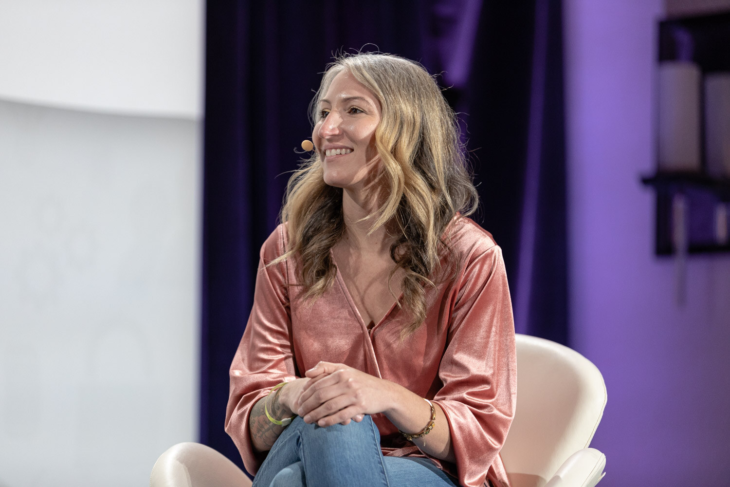 Woman with long wavy blonde hair in a pink satin blouse and jeans seated with her hands on her knee, smiling, in front of a purple curtain.