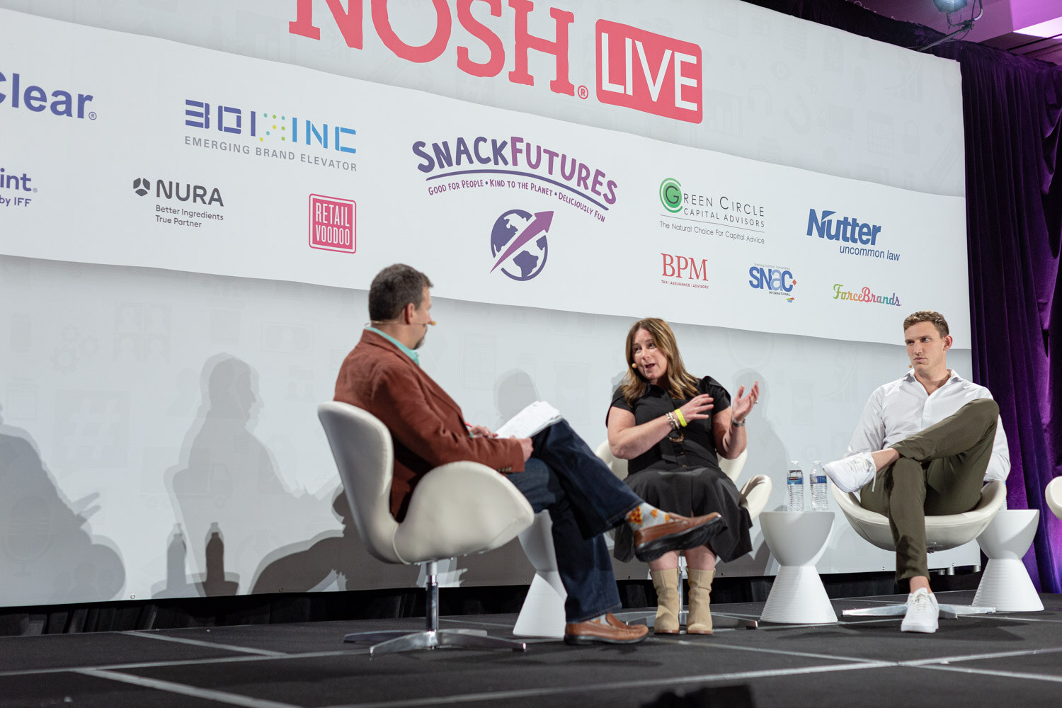 Three people seated in white chairs on stage in front of a Nosh Live banner with sponsor logos: a man in a rust jacket holding notes, a woman in a black dress and a man in a white shirt and olive pants.