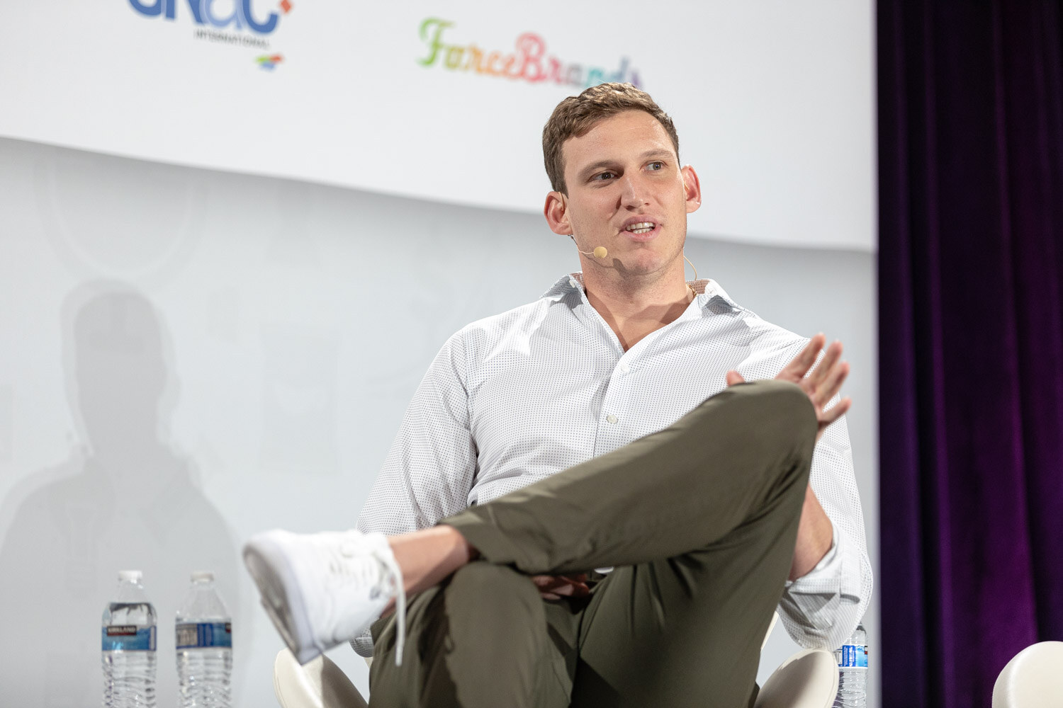 Man in a white button-up shirt and olive pants with one leg crossed over the other, gesturing with one hand, with two water bottles beside him.