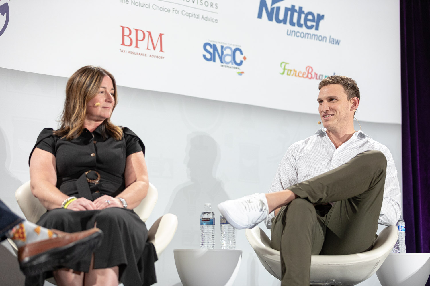 Woman in a black dress with a belt and a man in a white shirt and olive pants seated on white chairs in front of Nutter, BPM, SNAC and ForceBrands logos.