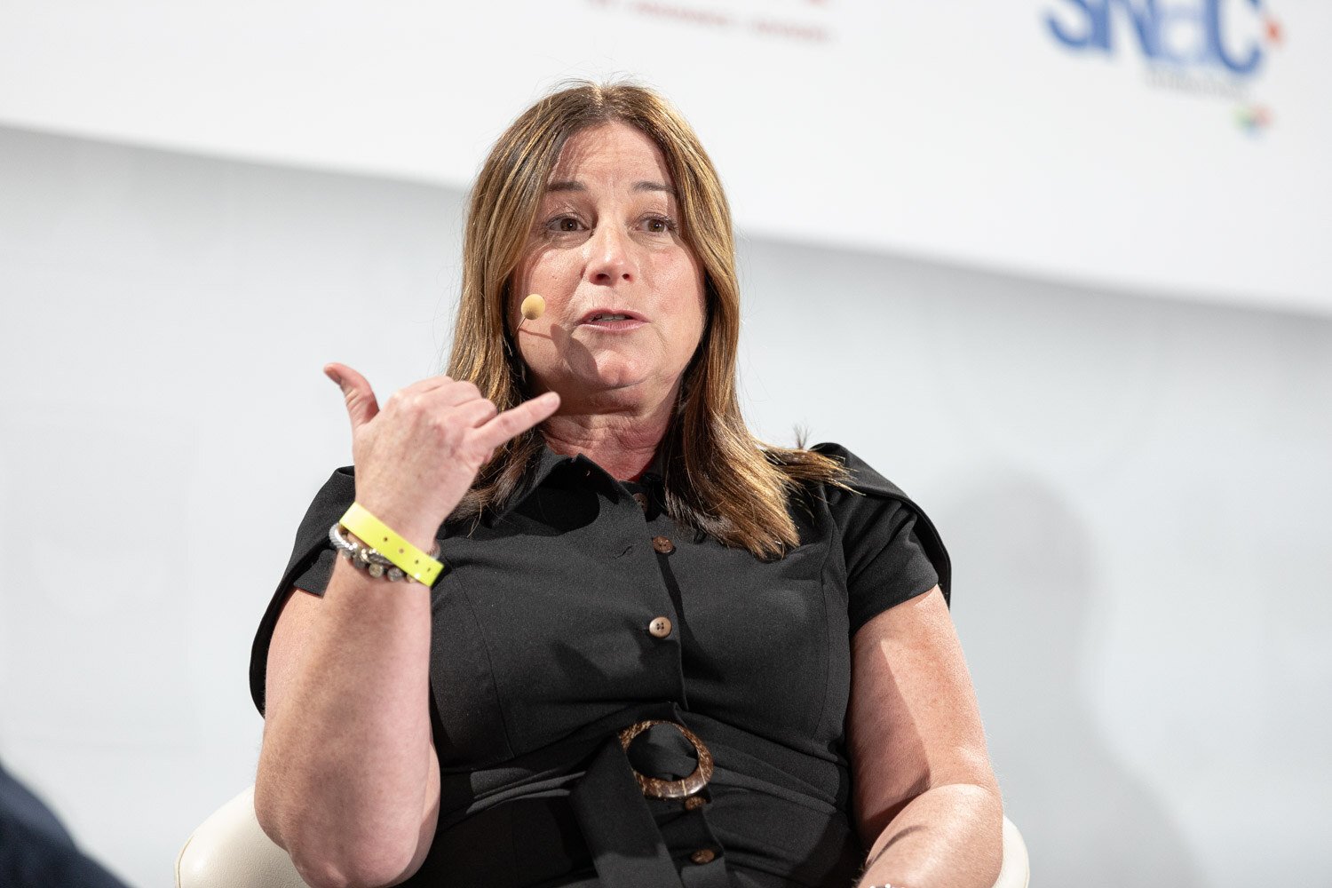 Woman with shoulder-length brown hair in a black short-sleeve belted dress and a yellow wristband making a gesture with one hand while speaking.