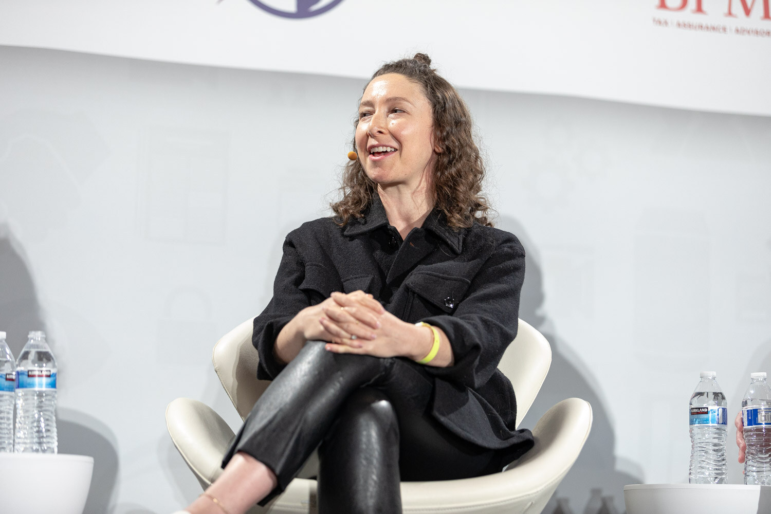 Woman with curly hair in a black overshirt and black leather pants seated with her hands around one knee, laughing, with water bottles on side tables.