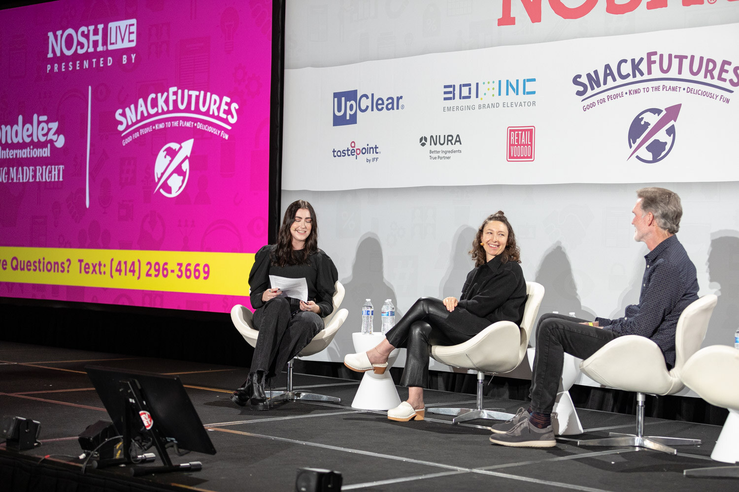 Three people seated on stage beside a large pink screen reading Nosh Live presented by Mondelez International and SnackFutures with a phone number for questions, and the sponsor wall at the right.