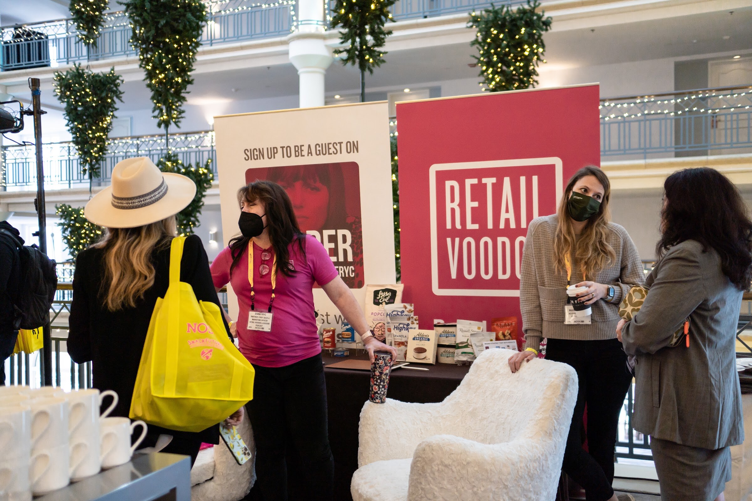 Attendees in masks and lanyards at a Retail Voodoo table with a white fur chair, with a woman in a hat carrying a yellow tote bag in the foreground and hanging lit garlands overhead.