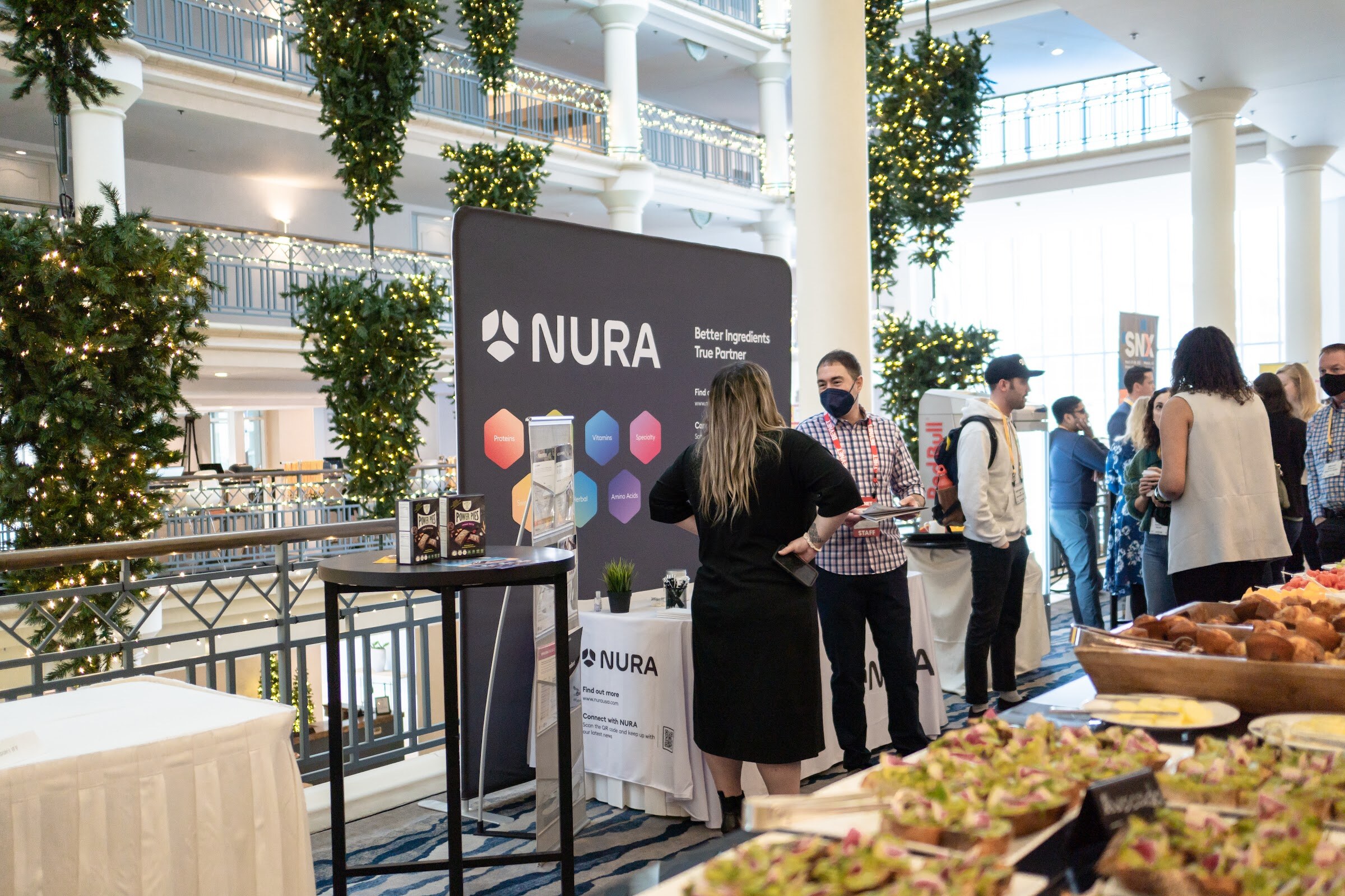 Dark NURA banner reading Better Ingredients True Partner on an atrium balcony with attendees in conversation, a table of pastries and avocado toast in the foreground and lit garlands overhead.