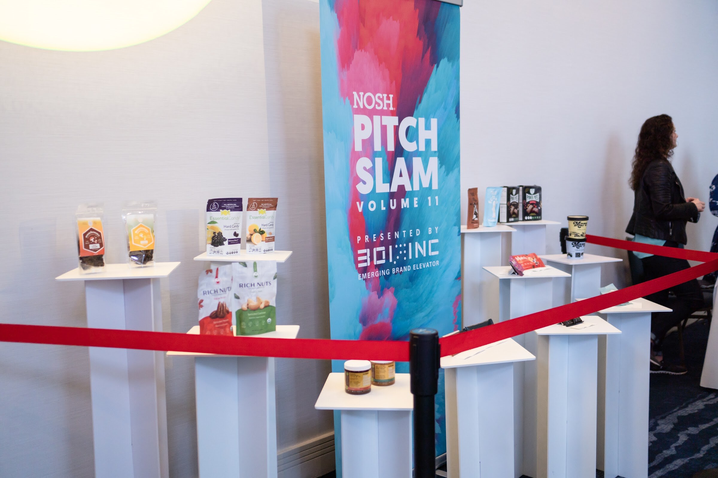 Banner reading Nosh Pitch Slam Volume 11 next to white pedestals holding Rich Nuts, EssentialCandy and Kokada packages and a red rope barrier.