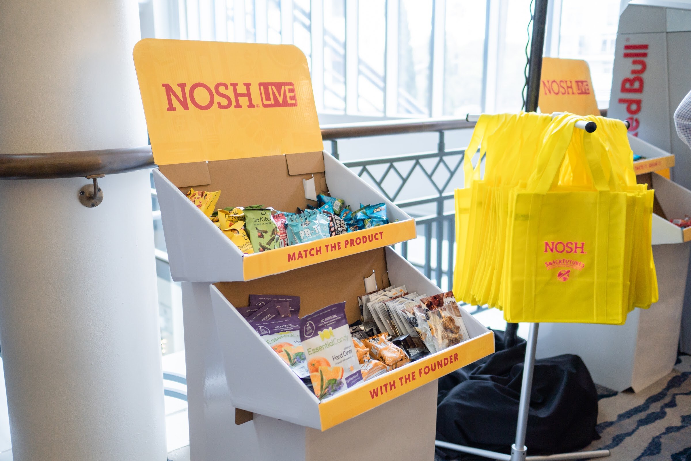 Yellow and white cardboard display reading Nosh Live with the words Match the Product with the Founder, holding snack packages, beside a stack of yellow Nosh tote bags.