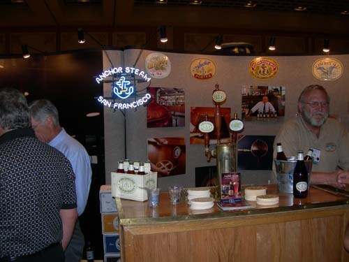 A bearded man in a tan shirt stands behind a wooden bar with taps and bottles under an Anchor Steam San Francisco neon sign, while two other men stand at left.