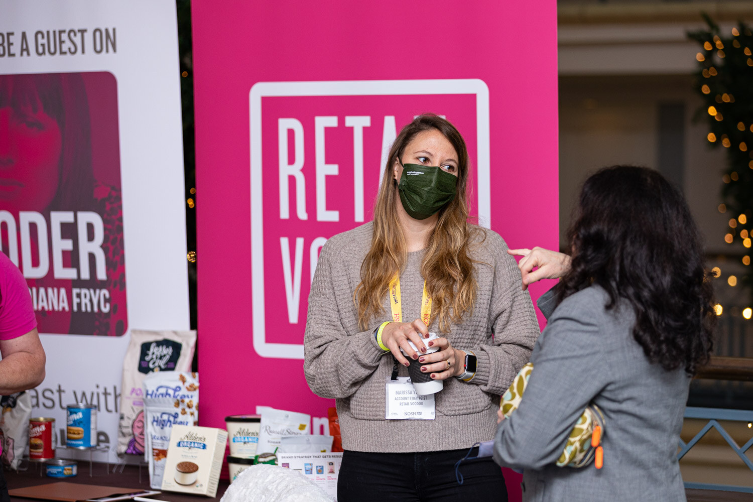 Woman in a green mask and tan sweater holding a coffee cup talking with a woman in a grey blazer, in front of a pink Retail Voodoo banner and a table of grocery products with a Sign Up to Be a Guest banner at the left.