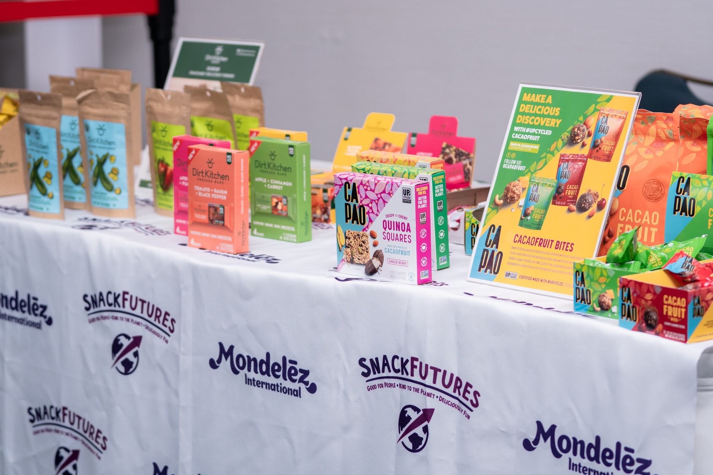 White tablecloth printed with Mondelez International and SnackFutures logos, holding Dirt Kitchen pressed bars, CaPao cacao fruit bites and quinoa squares in colorful packaging.