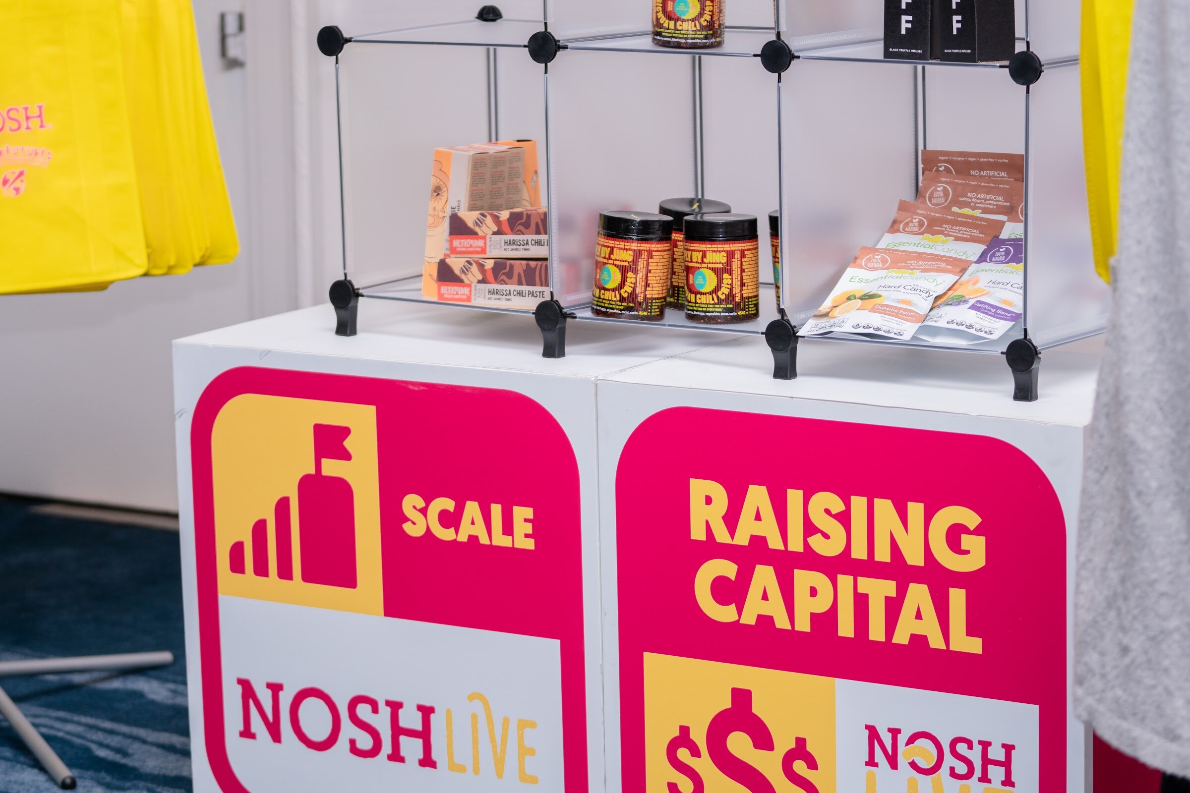 Pink and yellow display cubes reading Scale and Raising Capital with the Nosh Live logo, topped by a wire shelf of chili crisp jars and snack packages, with yellow tote bags at the left.