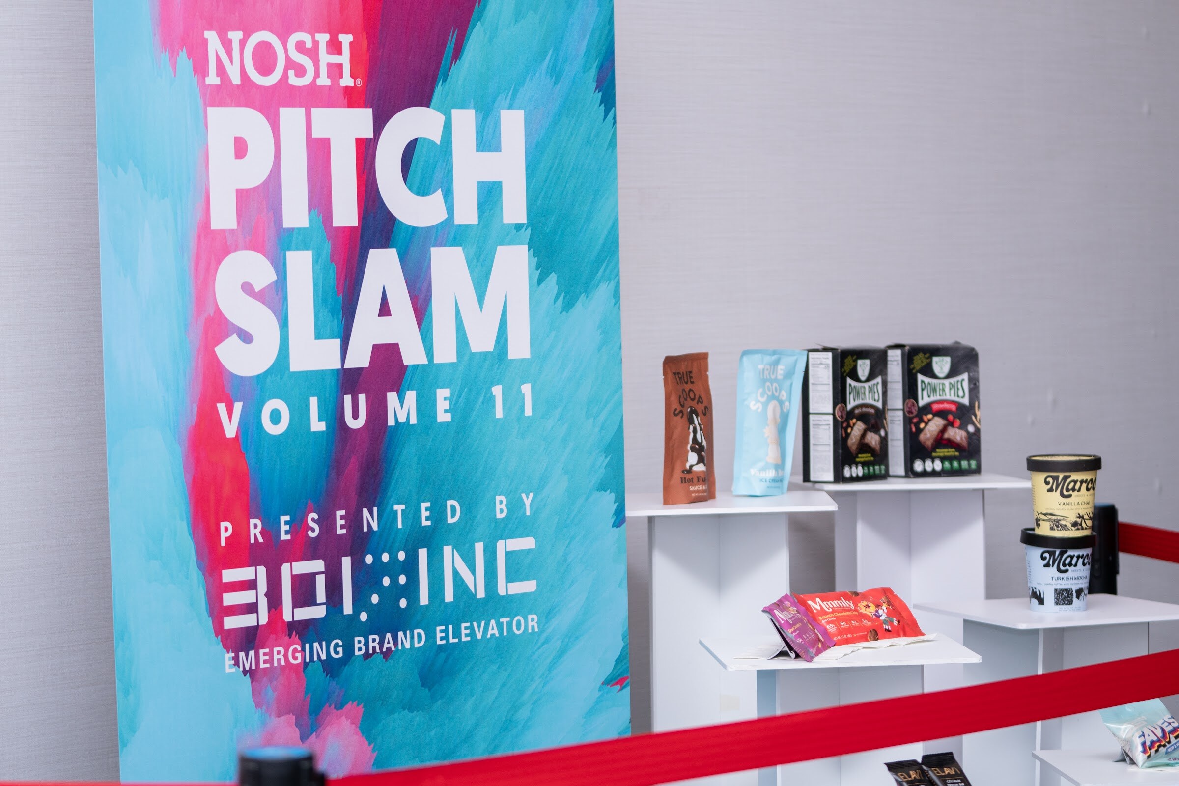 Banner reading Nosh Pitch Slam Volume 11 presented by 301 Inc Emerging Brand Elevator, next to white pedestals holding True Scoops, Power Pies, Marco ice cream pints and other snack packages.