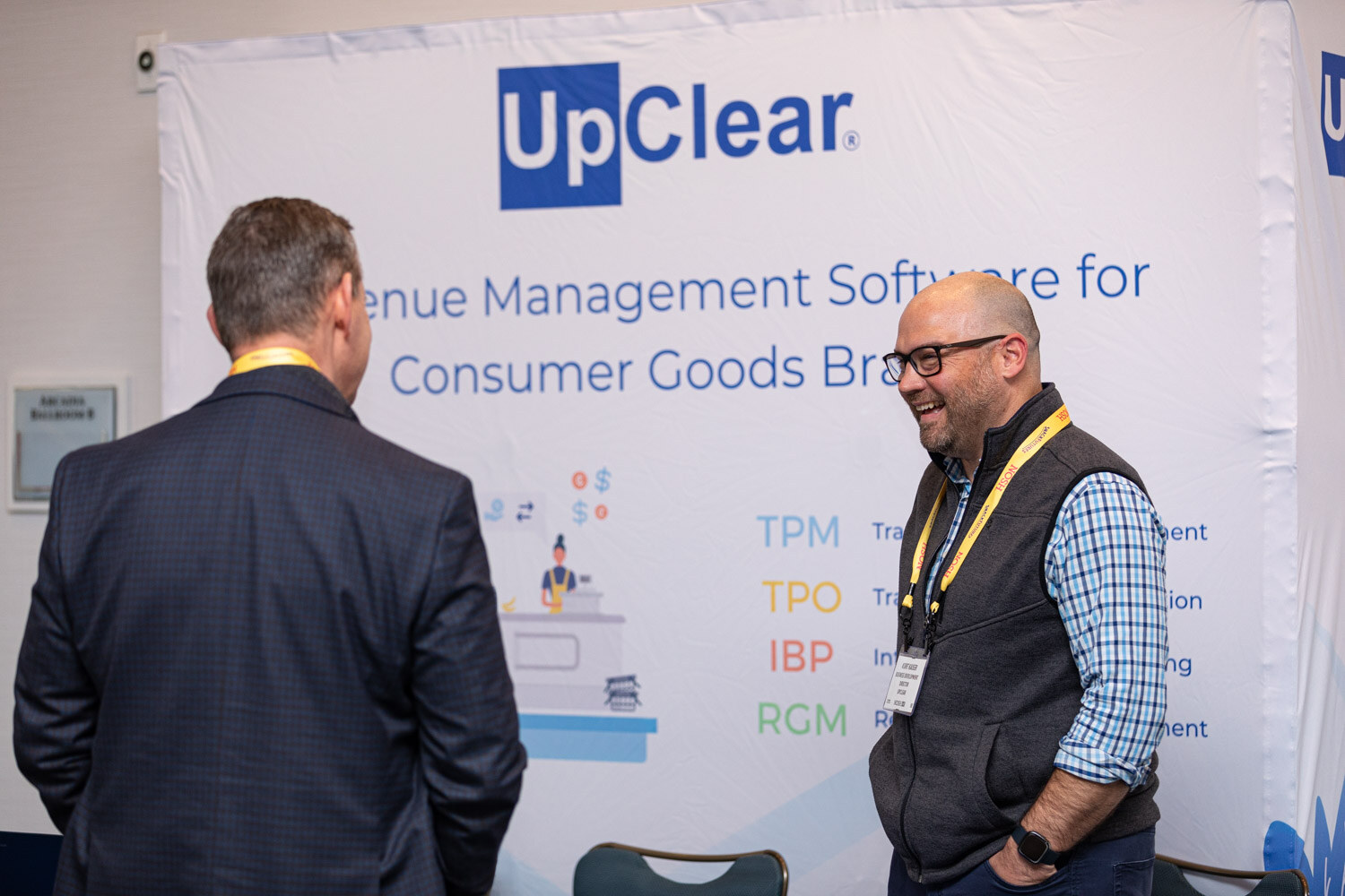 Man in a navy blazer seen from behind and a smiling man with glasses in a grey vest and checked shirt in front of a white banner reading UpClear Revenue Management Software for Consumer Goods Brands.