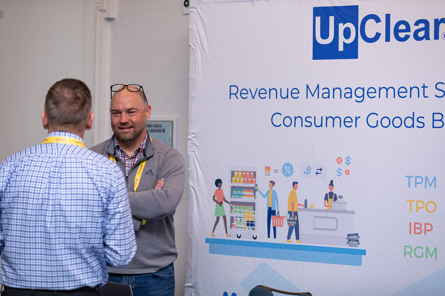 Man with glasses on his head and a grey fleece smiling and talking with a man in a blue checked shirt in front of an UpClear banner reading Revenue Management Software for Consumer Goods Brands.