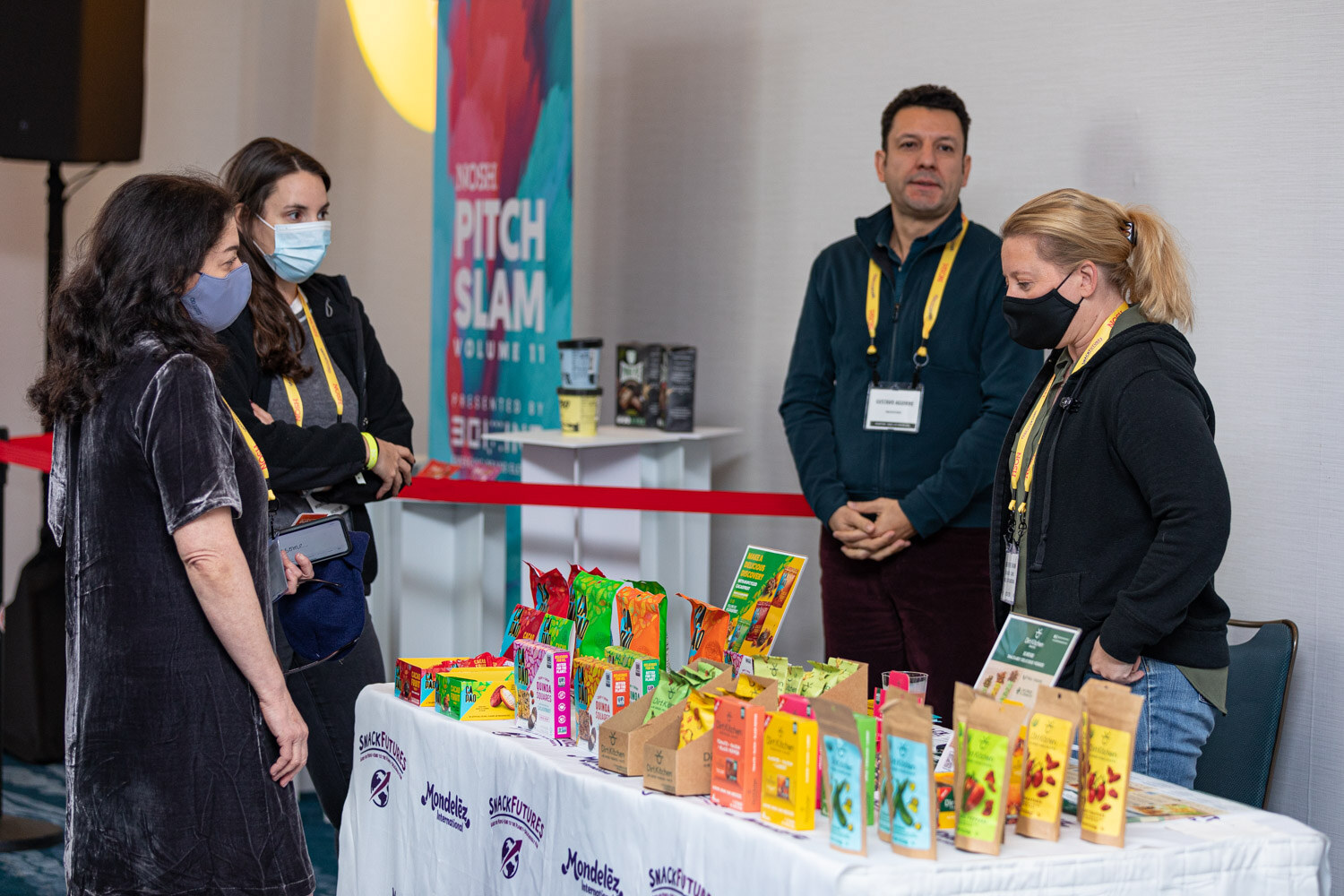 Four people with yellow Nosh lanyards at a white table printed with SnackFutures and Mondelez logos, covered with colorful snack packages, with a Nosh Pitch Slam Volume 11 banner behind.