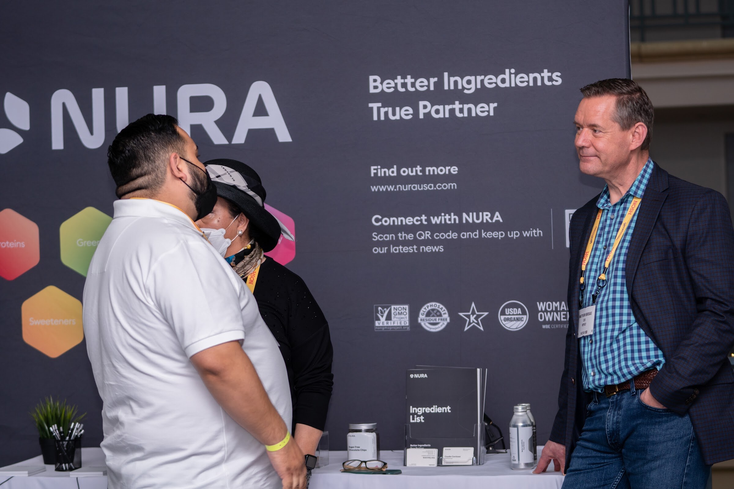 Man in a white polo and a woman in a hat and masks facing each other at the left and a man in a blue checked shirt and navy blazer at the right, in front of a NURA banner with certification logos.