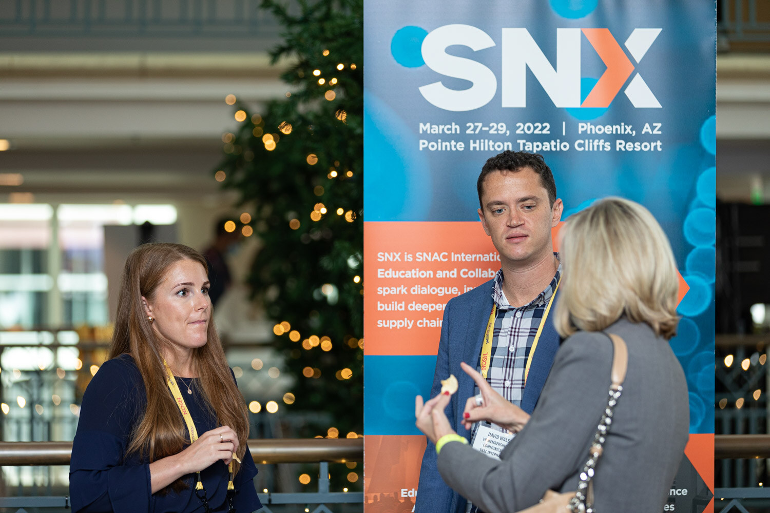 Man in a blue blazer and plaid shirt, a woman with long auburn hair and a woman in grey in front of a blue and orange SNX banner that lists March 27-29, 2022 at the Pointe Hilton Tapatio Cliffs Resort in Phoenix.