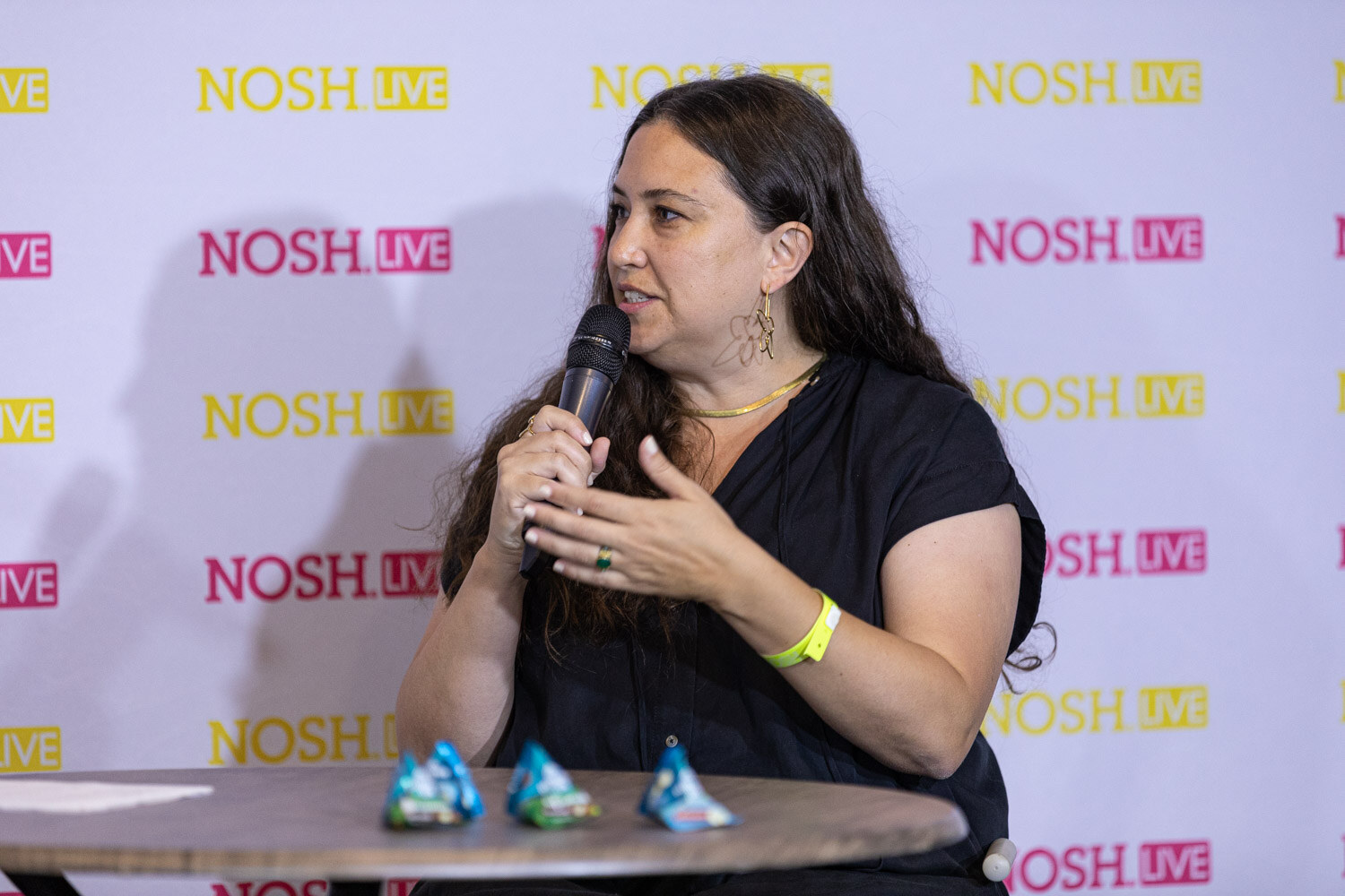 Woman with long dark hair in a black short-sleeve top and a gold necklace speaking into a microphone at a round wood table with three wrapped candies, in front of a pink and yellow Nosh Live backdrop.