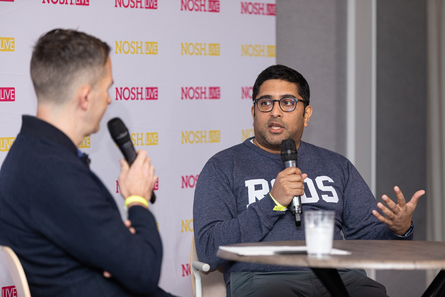 Man seen from behind in a navy sweater holding a microphone facing a man with glasses in a grey sweatshirt who gestures while speaking, at a round table in front of a Nosh Live backdrop.
