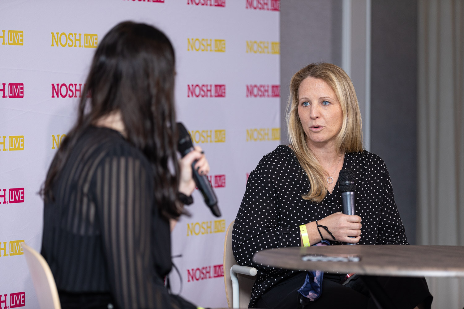 Woman with long dark hair seen from behind holding a microphone, facing a woman with blonde hair in a black polka-dot blouse holding a microphone at a round table, in front of a Nosh Live backdrop.