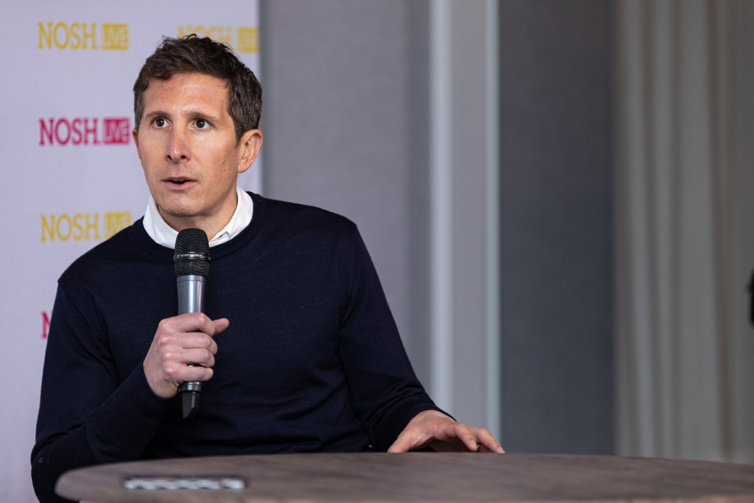 Man with short brown hair in a navy crew-neck sweater over a white collared shirt holding a microphone at a round table, in front of a Nosh Live backdrop and grey drapes.