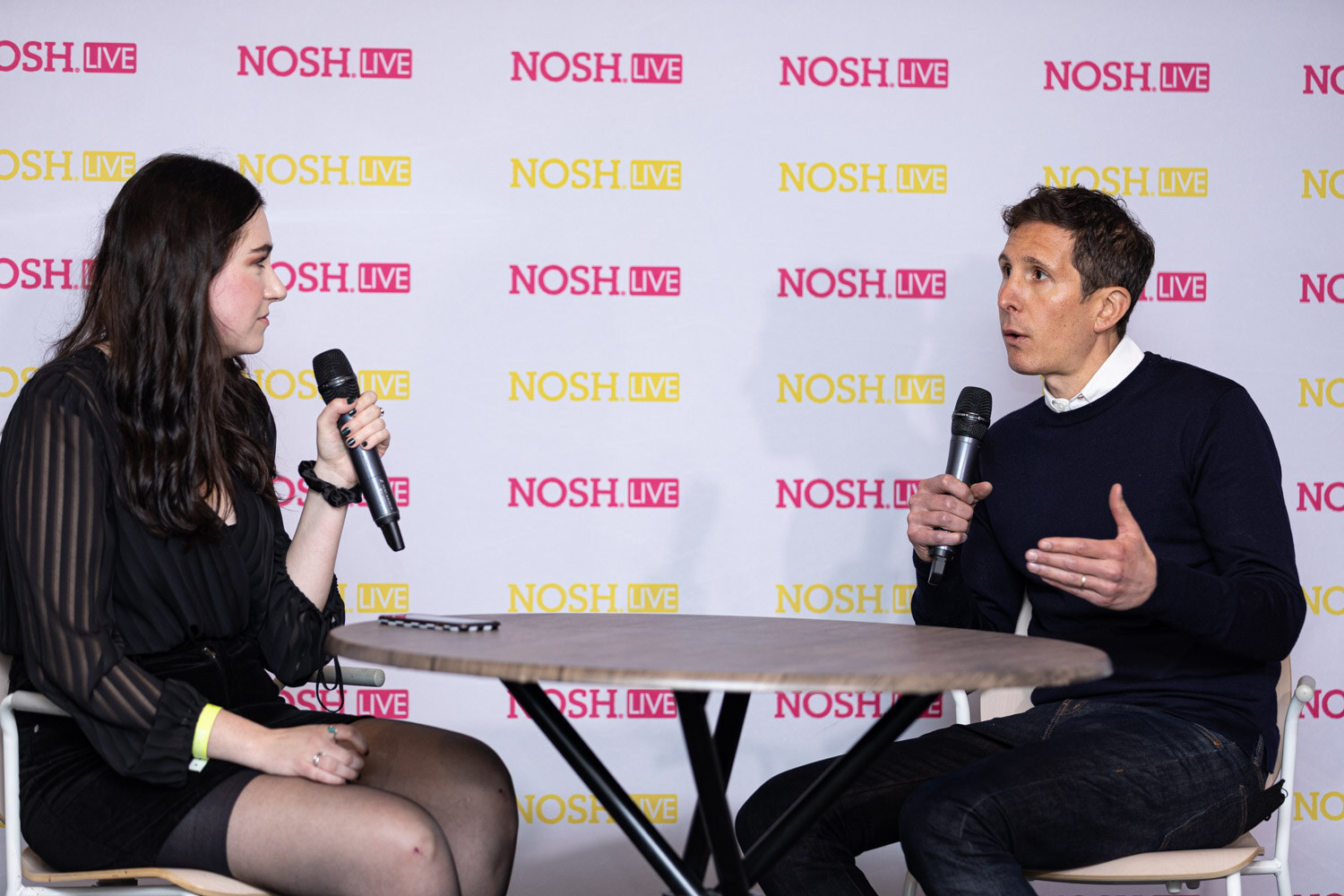 Woman with long dark hair in a black sheer top at the left and a man in a navy sweater at the right, both holding microphones at a round table in front of a Nosh Live step-and-repeat.
