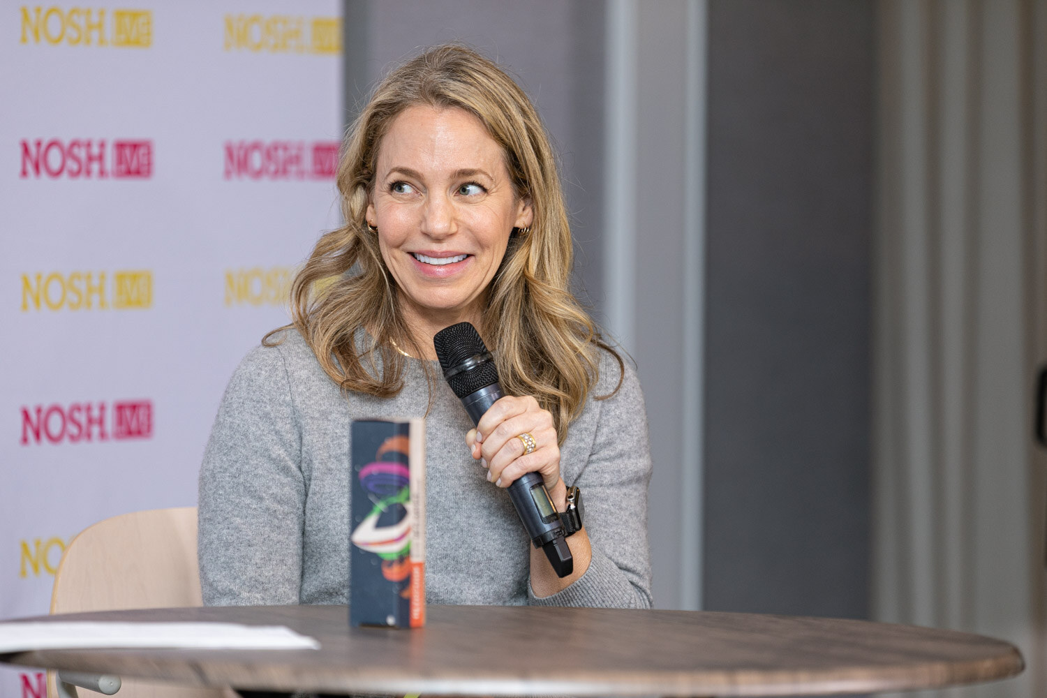 Woman with long wavy blonde hair in a grey sweater holding a microphone and smiling, with a book standing on the round table in front of her.