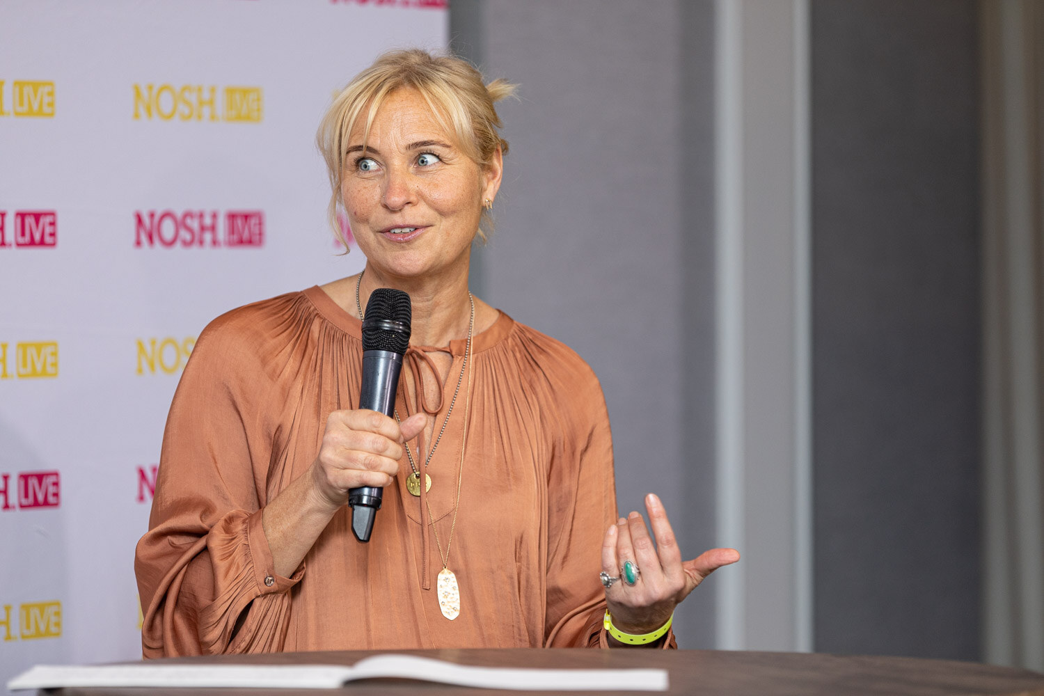 Woman with blonde hair in a bun and an orange satin blouse, with layered necklaces and a yellow wristband, holding a microphone and gesturing at a table with an open notebook.
