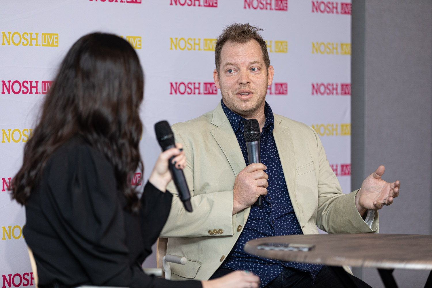 Man in a tan blazer and navy patterned shirt holding a microphone and gesturing with his right hand, seated at a round table across from the host.