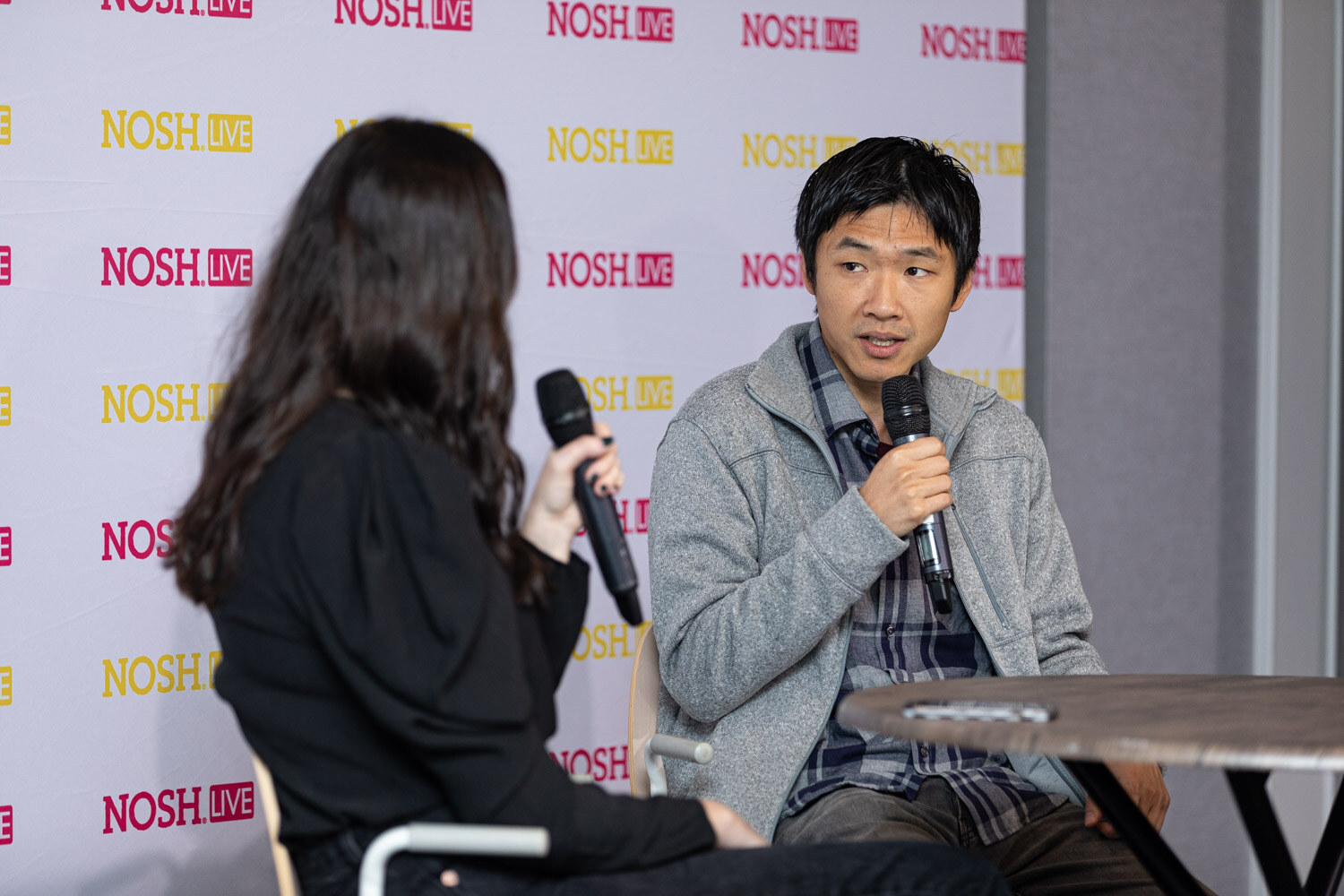 Man with short dark hair in a grey fleece jacket over a blue plaid shirt holding a microphone at a round table, with the host seen from behind at the left.