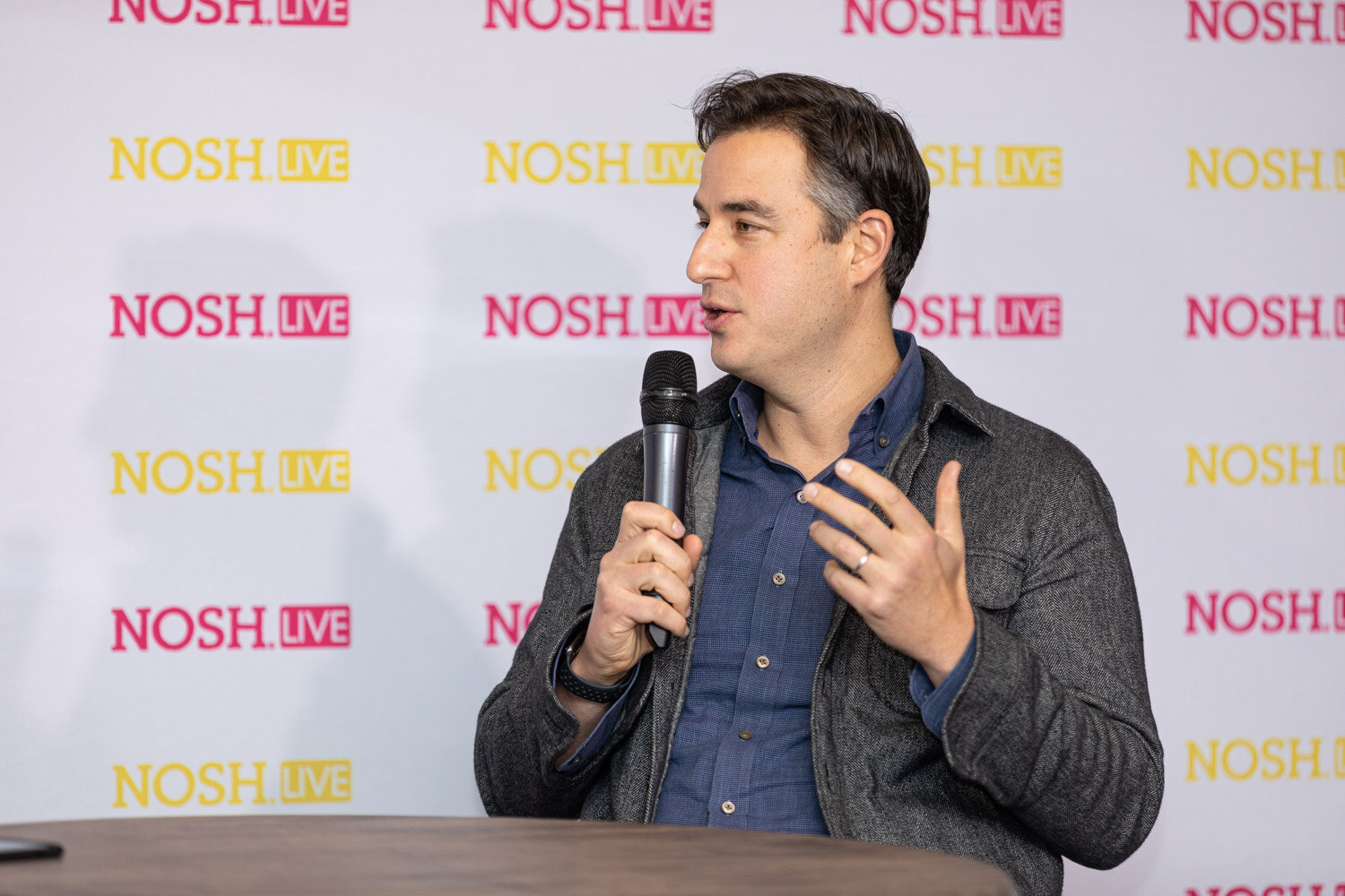 Man with short dark hair in a charcoal overshirt and blue button-up shirt holding a microphone and gesturing with one hand at a round table, in front of a repeating Nosh Live backdrop.