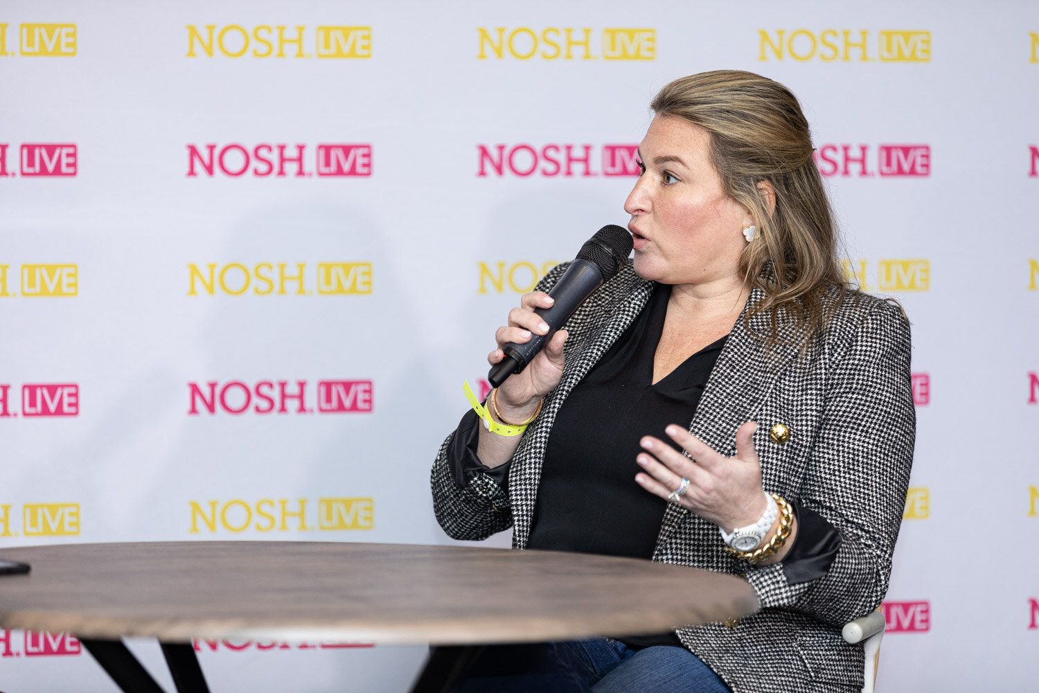 Woman with blonde hair pulled back in a black and white houndstooth blazer, black top and a yellow wristband speaking into a microphone in front of a Nosh Live backdrop.