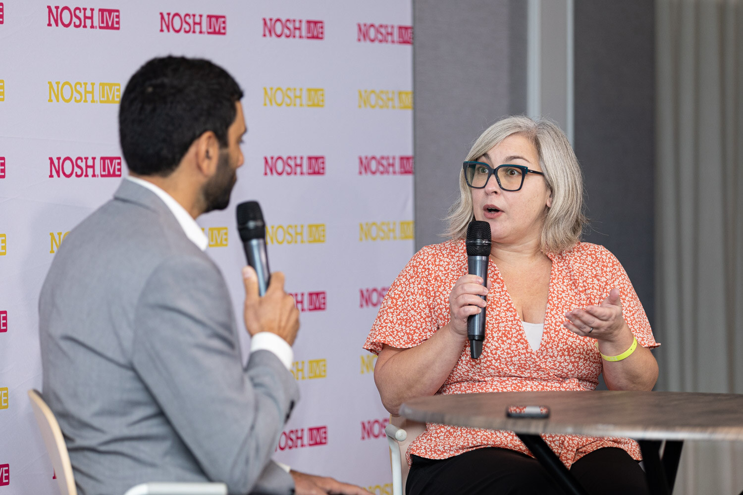 Woman with grey hair and dark glasses in an orange floral blouse holding a microphone and gesturing, facing a man in a grey blazer seen from behind at the left.