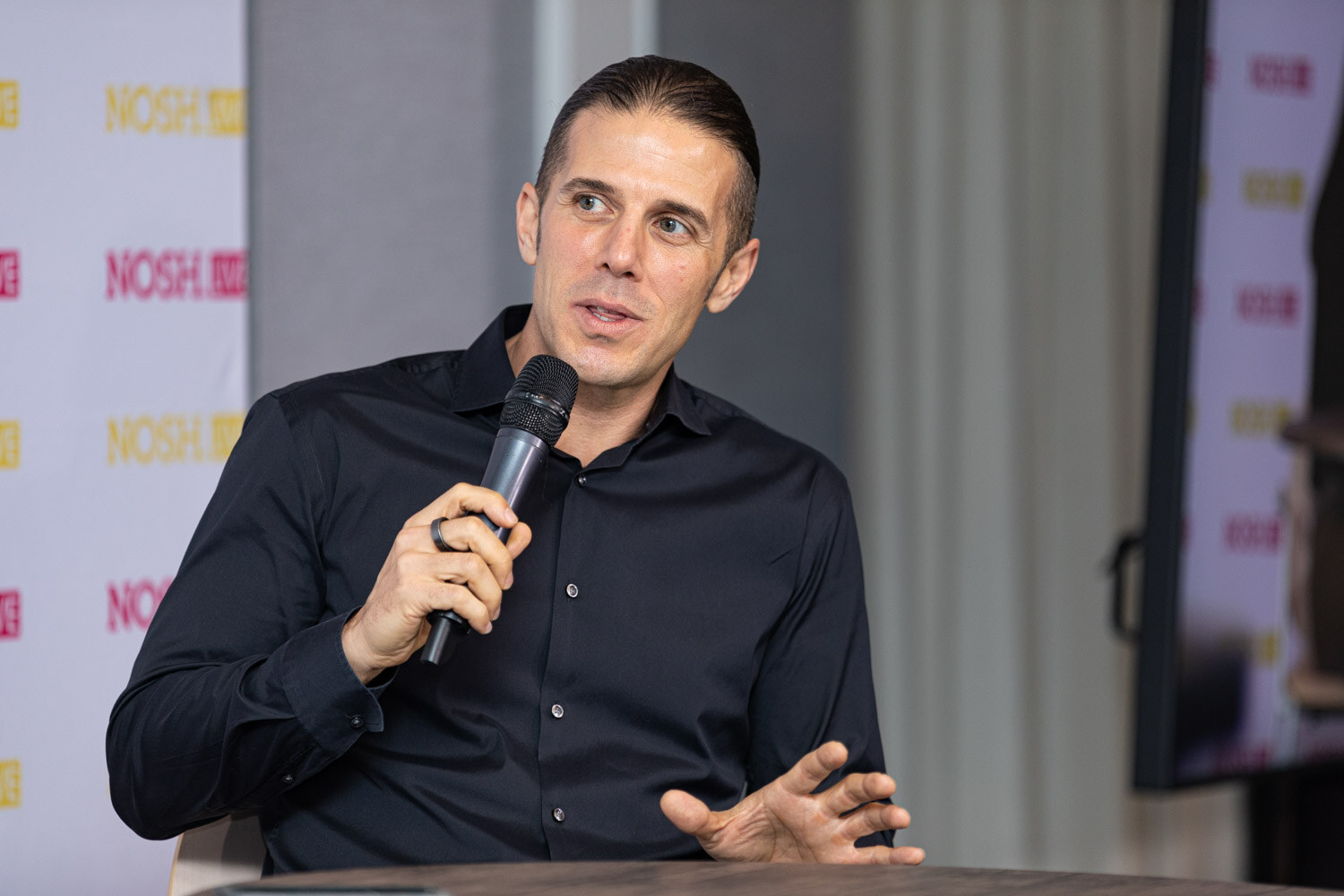 Man with slicked-back dark hair in a black button-up shirt holding a microphone in one hand and gesturing with the other, seated in front of grey drapes and a Nosh Live backdrop.