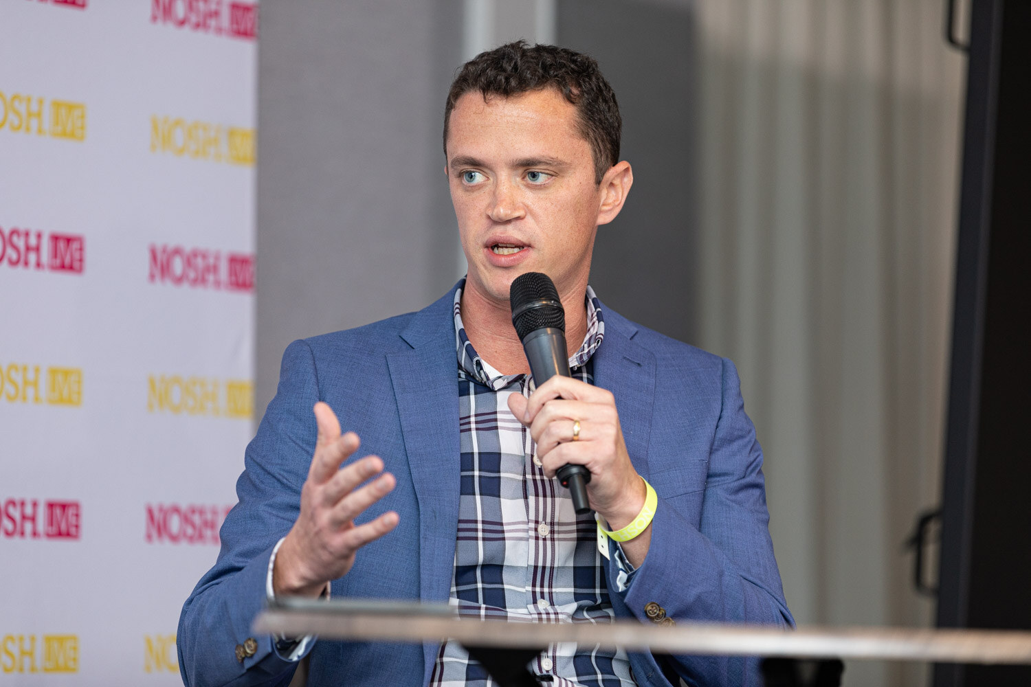 Man with short dark hair in a blue blazer over a navy and white plaid shirt holding a microphone and gesturing with one hand at a round table.